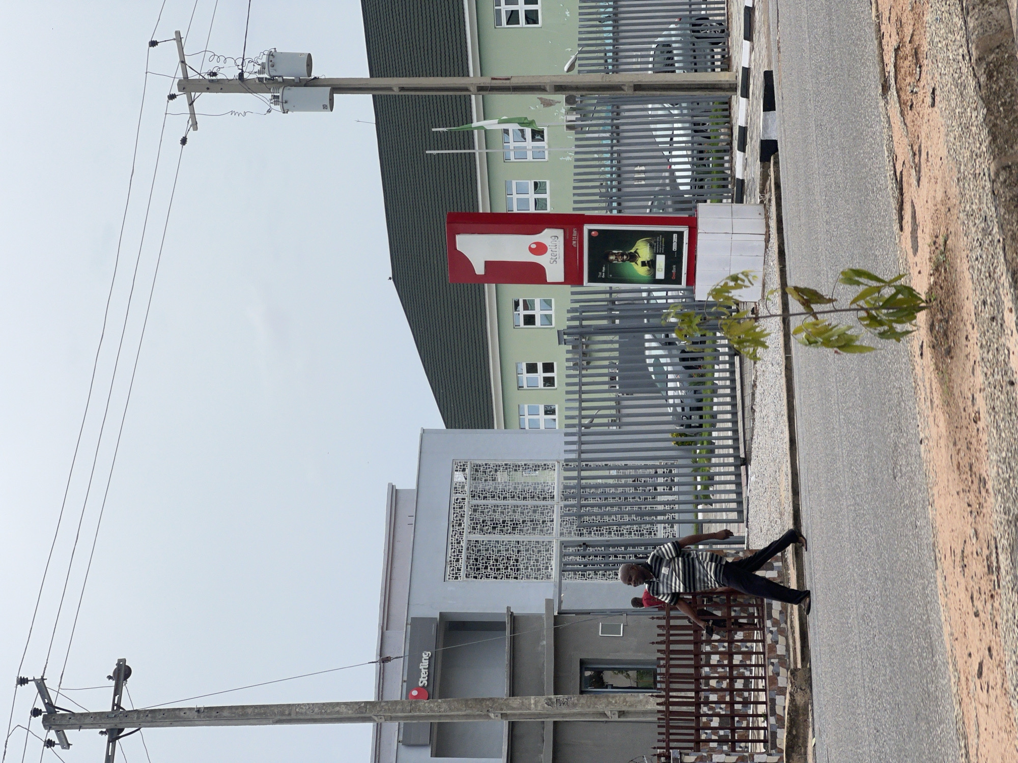 The image depicts an outdoor scene in Jalingo, Nigeria, featuring a Sterling Bank branch and its surrounding environment. A man in a striped shirt and dark pants is walking across the street in the foreground. Behind him, a gray metal fence encloses the bank property, which includes a modern, light gray building with distinctive patterned windows and a smaller building with a dark green roof. A large red sign with a white "1" and the Sterling logo stands prominently in front of the green-roofed building. Another sign below it displays a smiling man in a yellow shirt. A Nigerian flag on a flagpole is visible near the sign. Overhead, electrical wires are strung between concrete utility poles, one of which has transformers attached. A small, leafy tree grows on the roadside in the lower right corner. The sky is overcast.
