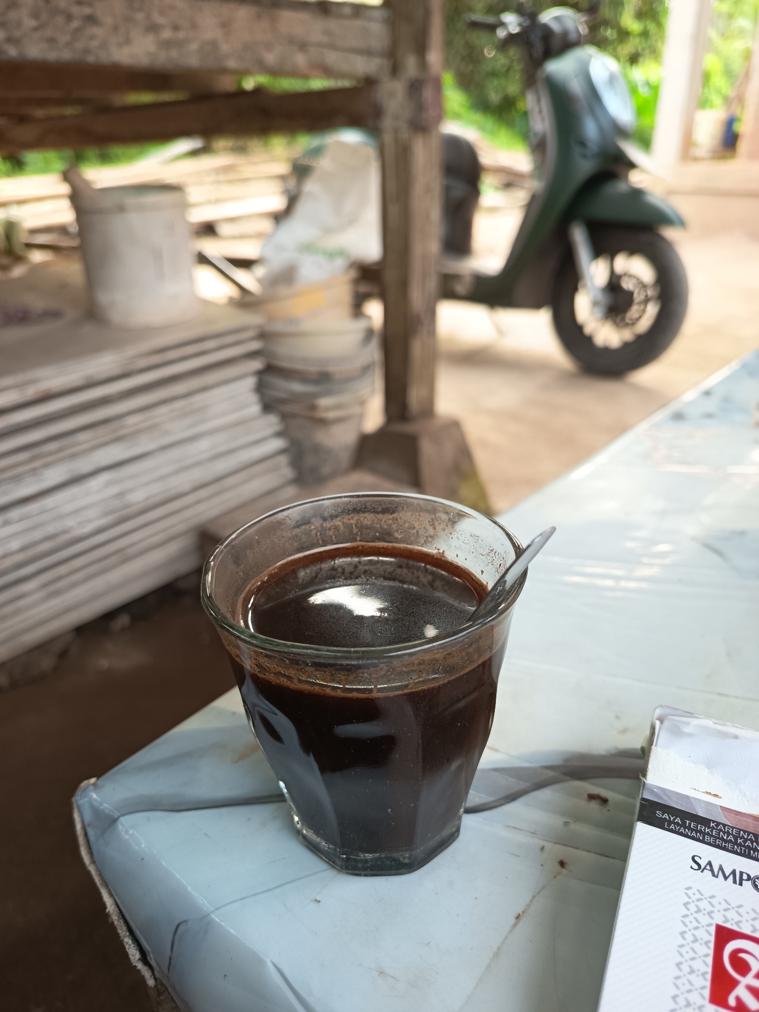 A glass of dark coffee sits on a light-colored, plastic-covered table in what appears to be an outdoor, covered area. A spoon is in the coffee. To the right of the coffee is a partially visible cigarette pack. In the background, a dark green scooter is parked. Behind the scooter, wooden planks are stacked, and buckets and bags are visible. The environment suggests a casual, possibly utilitarian setting, such as a workshop or a rural roadside stop. The lighting indicates it is daytime. The location is identified as Rayeukmatang, Indonesia.