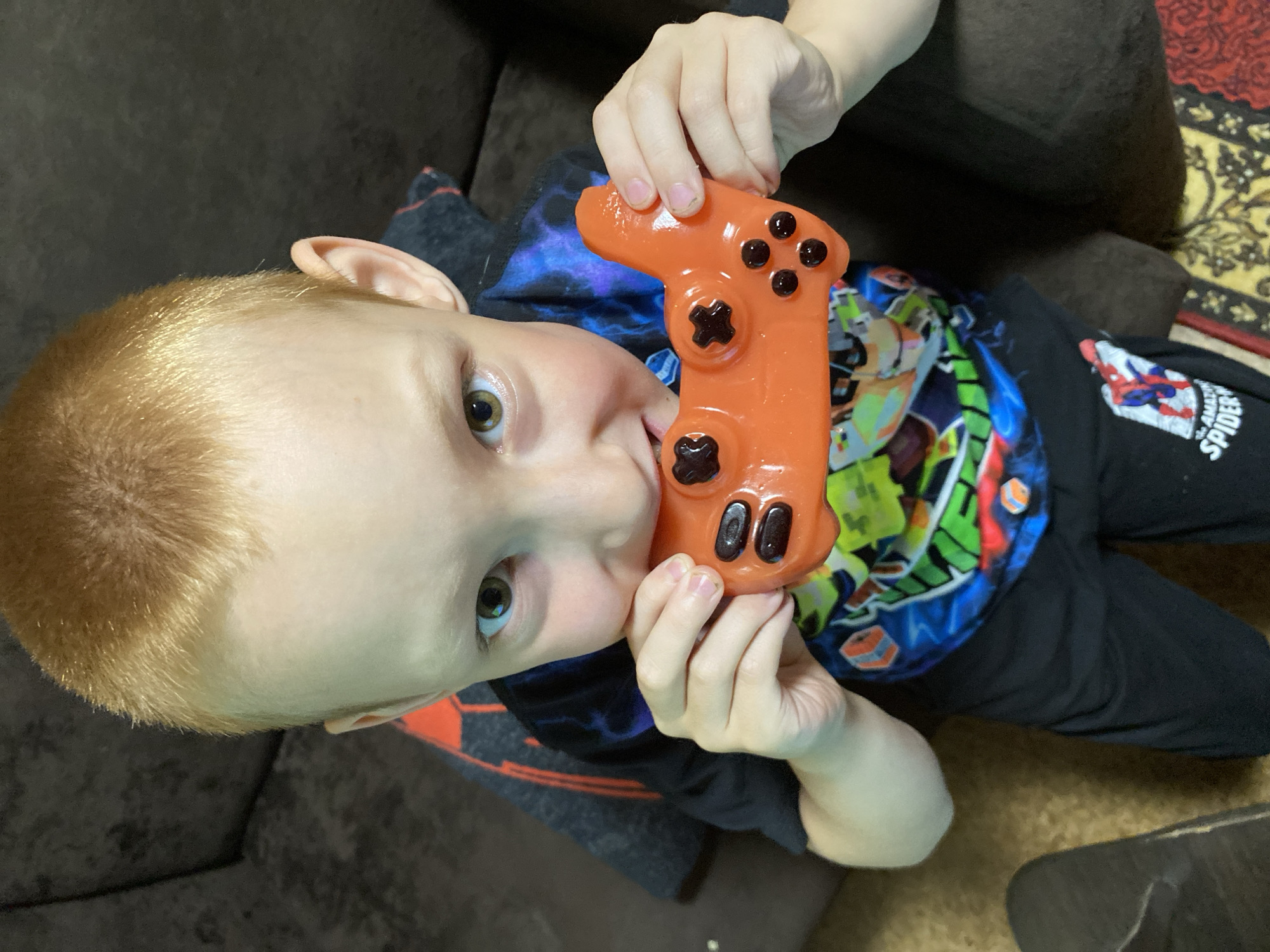 This media file features a close-up, high-angle shot of a young child with blond hair, holding an orange toy controller to their mouth. The child's eyes are wide and looking directly at the camera, with an expression that could be interpreted as playful or curious. The child is wearing a black t-shirt with a colorful graphic design, including what appears to be Spider-Man branding on the sleeve. The setting appears to be indoors, possibly on a couch, indicated by the dark fabric in the background and a patterned rug visible on the floor. The lighting suggests it is daytime. The toy controller is a bright orange and has black buttons. The overall mood of the image is candid and focuses on the child's interaction with the toy.