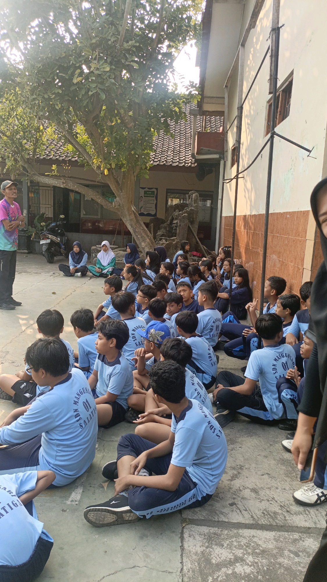 A group of students, mostly boys, are sitting on the ground in a schoolyard in Salatiga, Indonesia. They are all wearing light blue t-shirts with the text "SDN MANGUNSARI 01 SALATI" and dark blue pants or shorts. Some of the students are wearing headscarves. A man in a colorful t-shirt and a cap is standing and addressing the students. The school building is visible in the background, with a tiled roof and a light-colored wall. A large tree with green leaves is also present in the yard, casting shadows on the ground. The sun is shining brightly, indicating it is daytime. The scene appears to be an outdoor class or assembly.