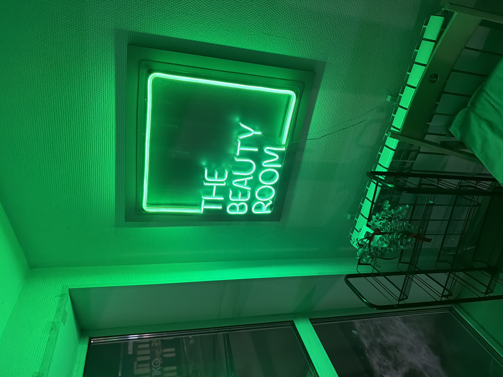 This image captures the interior of a commercial establishment, likely a beauty salon or similar service, in Chaikovsky, Russia. The scene is dominated by a pervasive green illumination originating primarily from a rectangular green neon sign mounted on a textured wall on the right side of the frame. The sign displays the text "THE BEAUTY ROOM" in capital letters. A large window on the left side of the room reflects external elements, including a partial view of a sign with Russian text "ТIКОМЕ 24," suggesting a commercial building or address visible from the street in Chaikovsky. Beneath the window, a dark, multi-tiered metal shelf holds a small artificial plant with green leaves. A white panel radiator is visible below the neon sign, and a portion of a light-colored chair or bench with a green cushion is present in the lower right foreground. No individuals are present, and the entire room, including its objects and surfaces, is bathed in a distinct green hue.