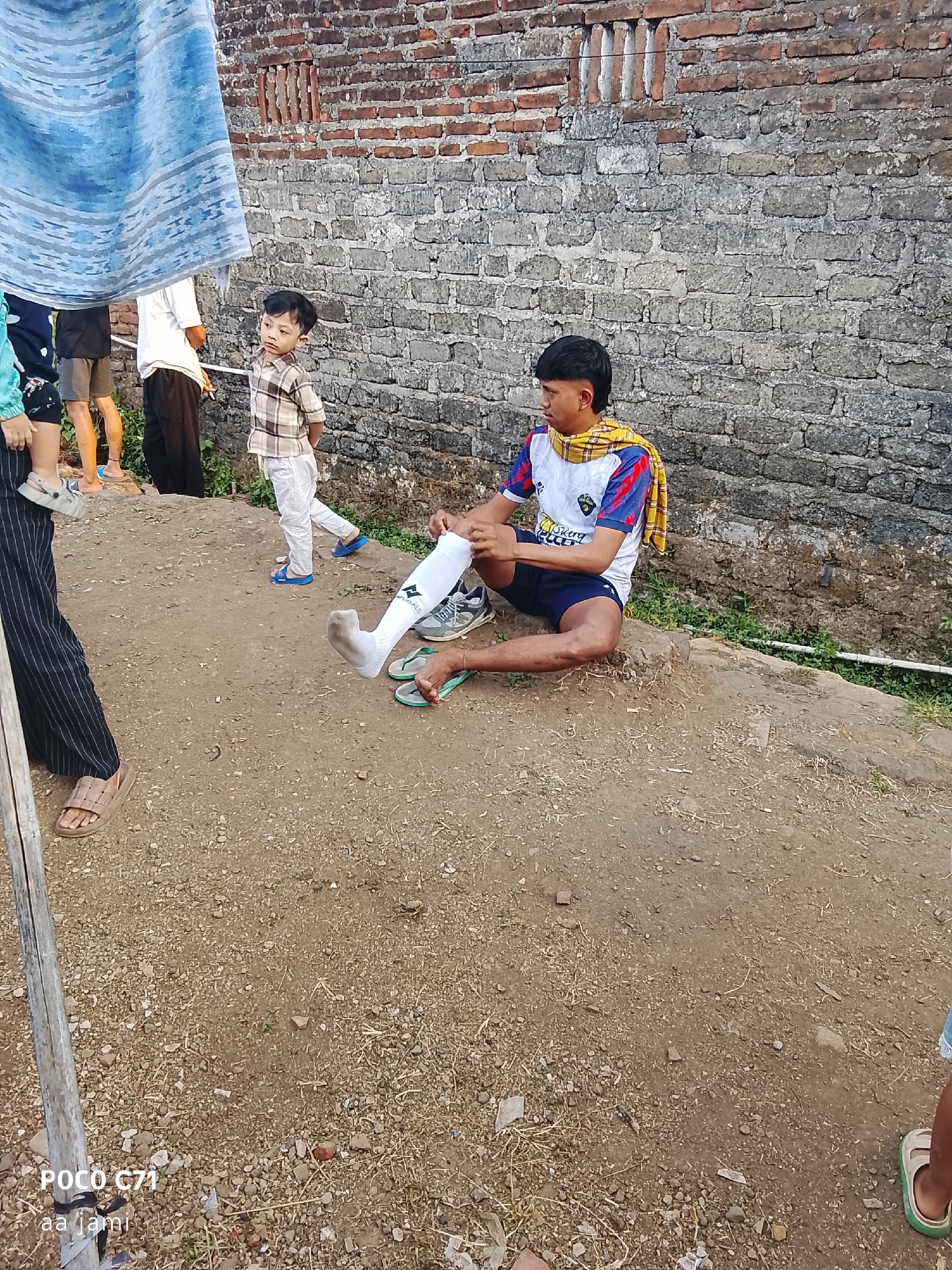 A low-angle, eye-level medium shot shows several people in an outdoor, dusty environment in front of a brick wall in Tasikmalaya, Indonesia. The scene is partially obscured by a blue and white striped cloth hanging from the upper left corner. In the foreground, a young man is sitting on a dirt mound, pulling a white sock onto his right foot, which is resting on a grey sneaker. He wears a white shirt with red and blue sleeves, blue shorts, and a yellow and white checkered scarf around his neck. To his left, a younger child in a checkered shirt and white pants stands looking to the right, with hands clasped behind his back. A portion of another person's legs wearing black and white striped pants and brown sandals is visible on the far left. Behind the child, another person in a white shirt and dark pants stands. A wall made of weathered bricks forms the backdrop, with some sparse green vegetation growing at its base. The ground is covered in dirt, small stones, and dry grass. The lighting suggests it is daytime.