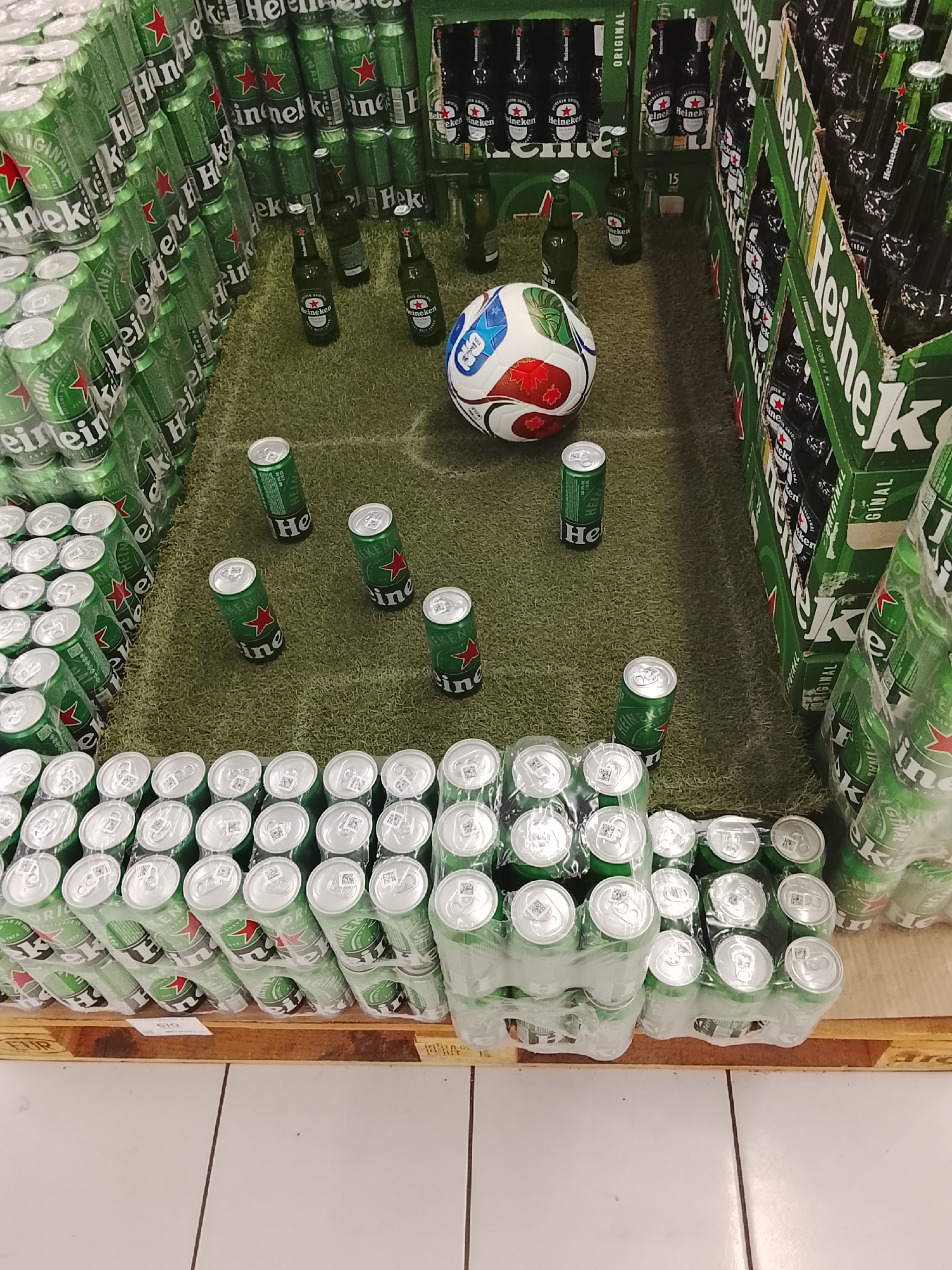 The image shows a retail display of Heineken beer products arranged to resemble a soccer field. Numerous green Heineken cans and bottles are stacked in rows, with a soccer ball placed in the center of the arrangement. The display is situated on a green surface that mimics grass, complete with white markings to simulate field lines. In the background, additional cases of Heineken beer are visible. The scene appears to be indoors, possibly in a supermarket or a beverage store. There are no people visible in the image.