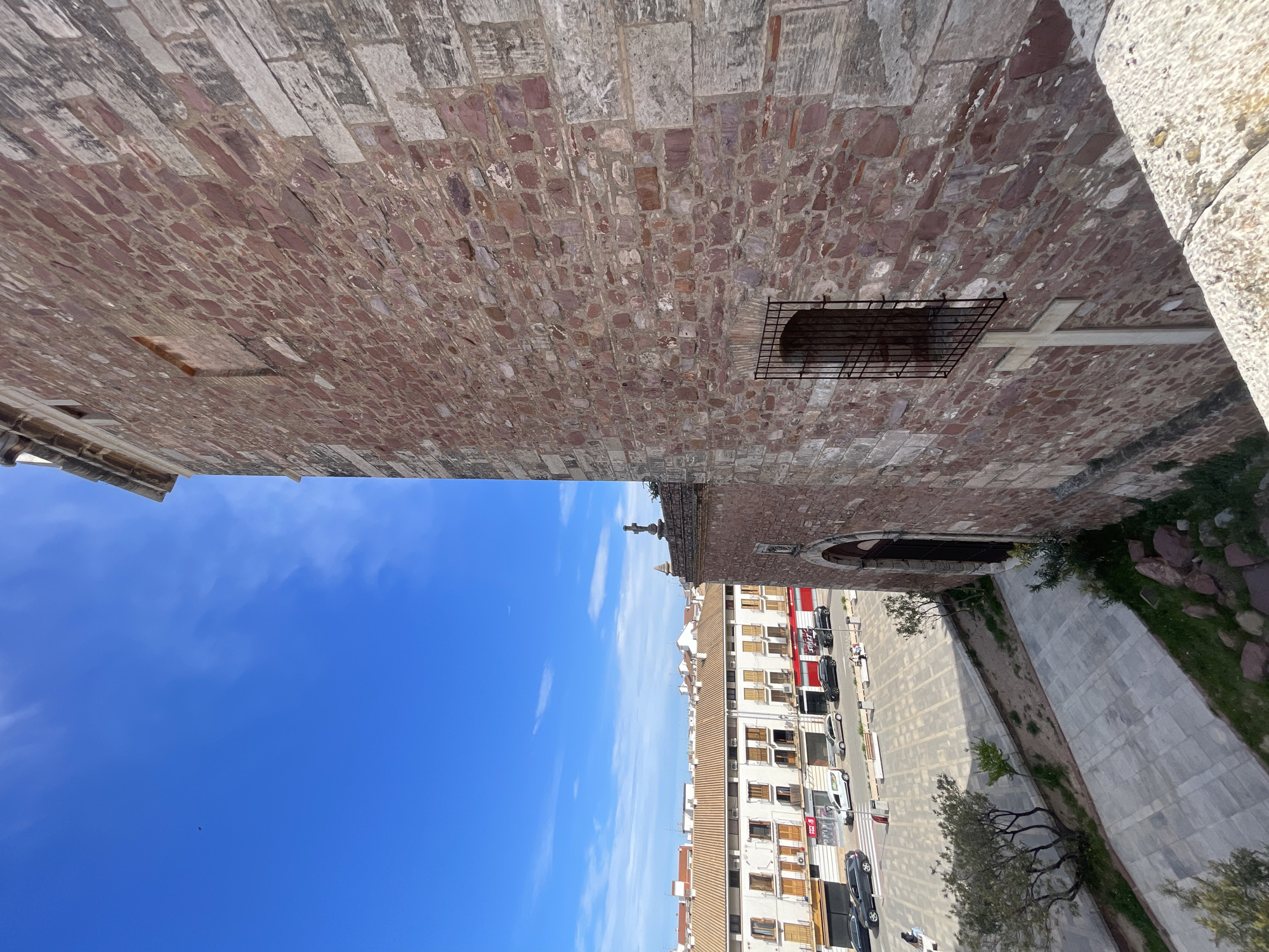 The image depicts an upward-facing view in El Puig De Santa Maria, Spain, revealing an old stone building structure contrasting with the bright blue sky. The architecture suggests historical significance with its aged bricks, a barred window, and a cross symbol integrated into the design. Below, a portion of the city is visible, with multi-story buildings lining a street, marked by parked cars and hints of commercial activity. Vegetation creeps along the building's base, adding a touch of nature to the urban landscape. The composition emphasizes the juxtaposition of the historical building against the modern city backdrop under a clear, daytime sky. There are no people visible in the frame.