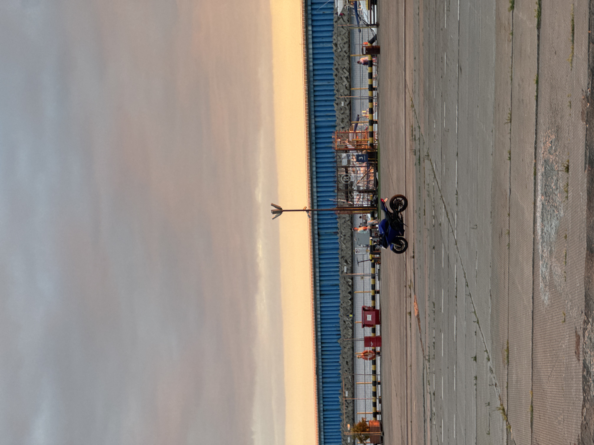 A blue motorcycle is parked in an open, paved area, possibly a port or dock. In the background, a long, blue structure runs horizontally across the image, topped with a railing. The sky above is filled with clouds, with a gradient of orange and pink hues suggesting either sunrise or sunset. Several people are visible in the background near the blue structure, and a few objects like red bins and a life preserver are scattered along a yellow and black striped barrier. The paved area in the foreground has some weeds growing through cracks. The scene appears to be in Sirius, Russia, likely a public area near the waterfront.