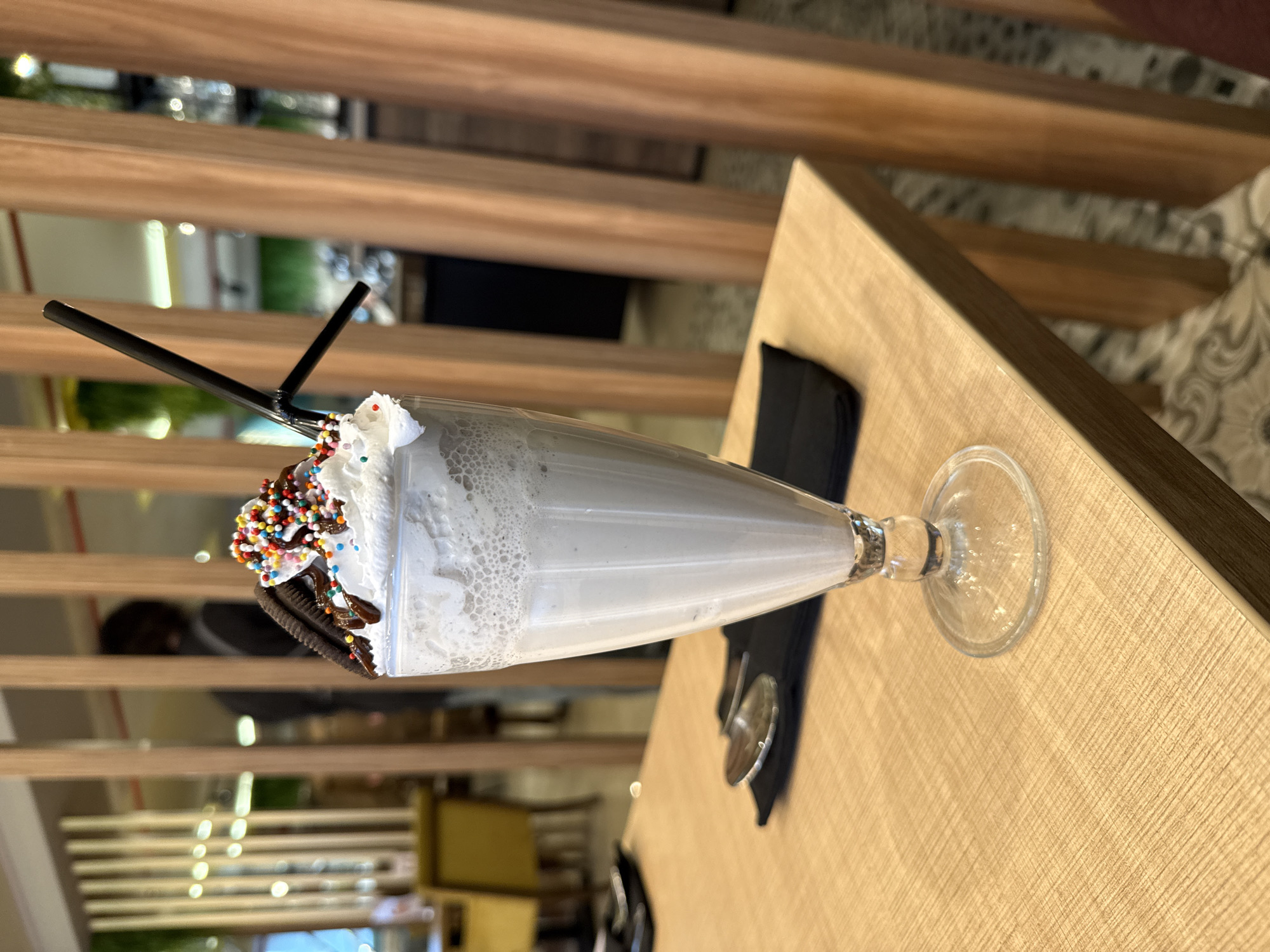 A milkshake rests on a wooden table in what appears to be a restaurant. The milkshake is in a clear glass with a stemmed base. It is topped with whipped cream, chocolate syrup, sprinkles, and two cookies. Two black straws are inserted into the milkshake. A black napkin and silver utensils are on the table beside the milkshake. The table is light brown with a textured surface. In the background, there is a wooden slat wall and glimpses of the restaurant's interior. The location is not determined.