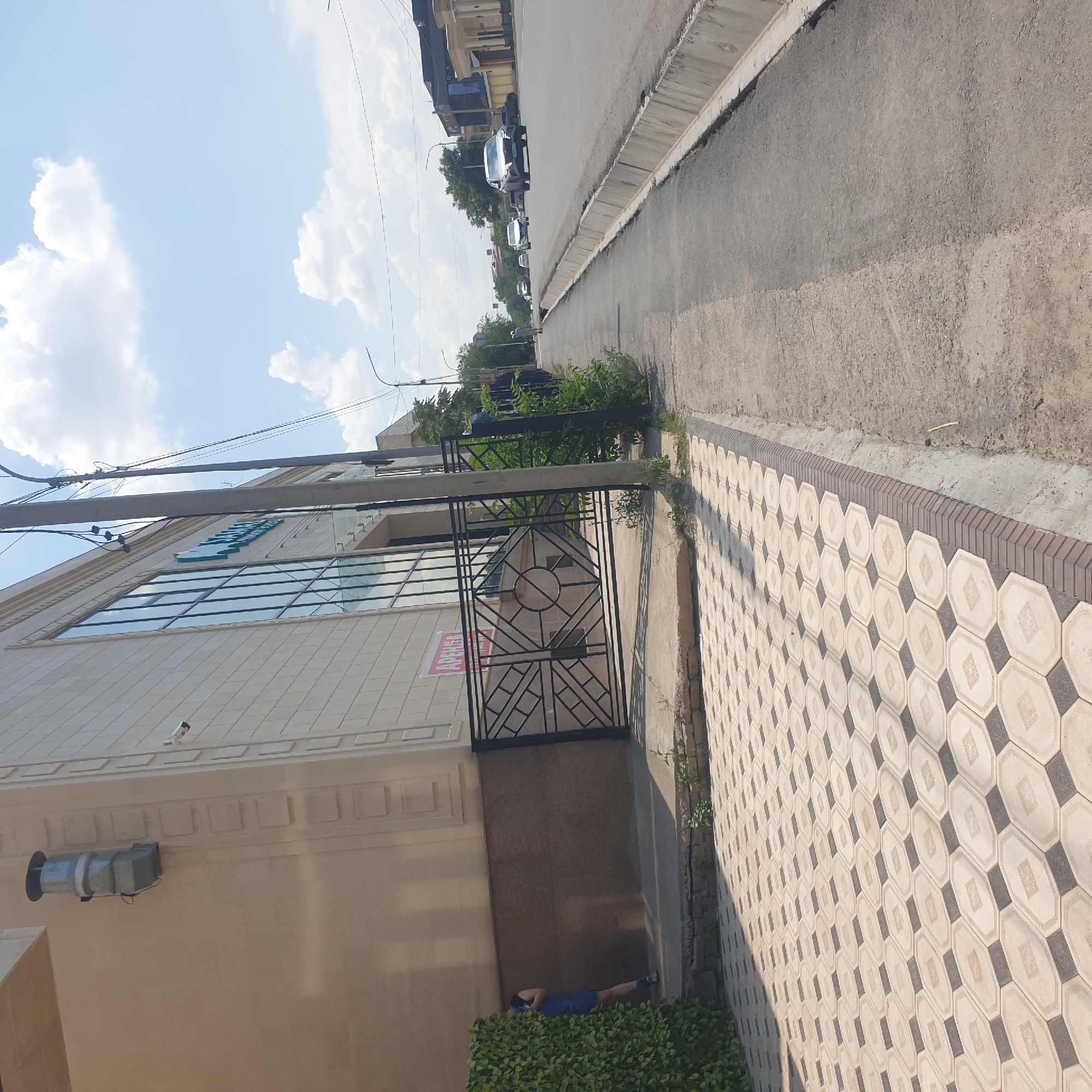 A bright, sunny day is depicted on a city street in Shymkent, Kazakhstan. In the foreground, a patterned tiled walkway extends from the left side of the frame, transitioning to a concrete sidewalk. A dark green hedge is visible on the far left. Adjacent to the walkway is the beige facade of a modern building with large glass windows. A black metal fence with an intricate geometric design stands before the building entrance, partially obscuring a red "АРЕНДА" (rent) sign. To the right of the building, a grey concrete pole with wires runs vertically. A person in a blue dress is partially visible behind the hedge on the left. The street itself is paved, with a concrete drainage ditch running along the right side. Several cars are traveling on the street, including a silver SUV in the foreground and other vehicles further down the road. Trees and other buildings line the far side of the street, under a blue sky with scattered white clouds. A security camera is mounted high on the building's facade.