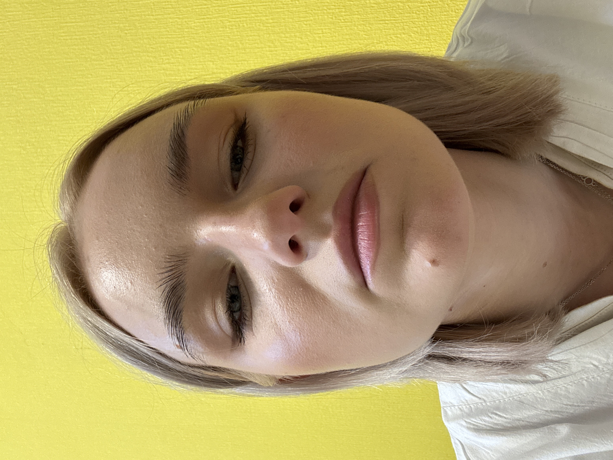 A close-up portrait features a young woman with light, possibly ash blonde hair, set against a vibrant yellow, textured background. Her head is tilted slightly back, and she is looking downwards with her eyes partially closed, giving her a serene or pensive expression. She has prominent dark eyebrows and full lips with a subtle pink hue. Her skin appears smooth with a slight sheen, and a small mole is visible below her right lip.
She is wearing a light-colored, likely white or off-white, collared shirt, visible on the right side of the frame, and a thin silver chain is around her neck. The lighting is soft and even, gently illuminating her features. No specific activity is taking place beyond posing for the portrait. The background is a plain yellow wall, suggesting an indoor setting. There are no visible cues for the time of day, weather, or specific location, other than the general context of Russia. No text is visible in the image.