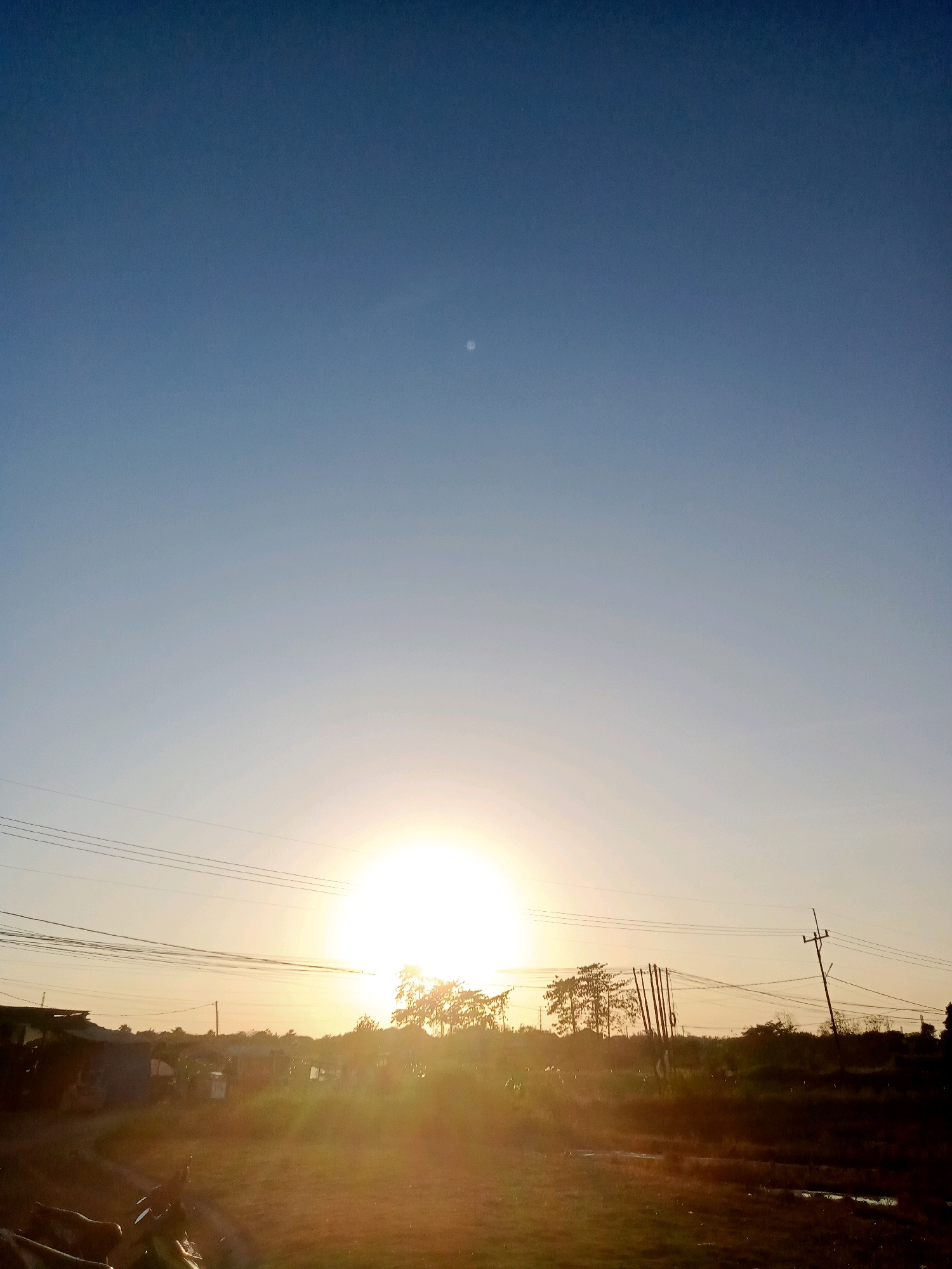 This image captures a serene sunset over a rural landscape in Banjarwangunan, Indonesia. The sky transitions from a deep blue at the top to a soft gradient of pale yellow and orange near the horizon, indicating the sun's descent. A small, pale celestial body, possibly the moon, is faintly visible in the upper portion of the sky.
The foreground is dominated by an unpaved road, with some debris and puddles suggesting recent rain or irrigation. Silhouetted against the bright sunset are trees and vegetation, some appearing to be cultivated. Several power lines stretch across the scene, supported by utility poles, indicating the presence of electricity infrastructure. In the mid-ground, there are hints of buildings and structures, suggesting a small settlement or village within the rural setting. The overall atmosphere is tranquil and natural, with the warm light of the setting sun casting long shadows and bathing the landscape in a golden hue. There are no people or animals directly visible in the image.