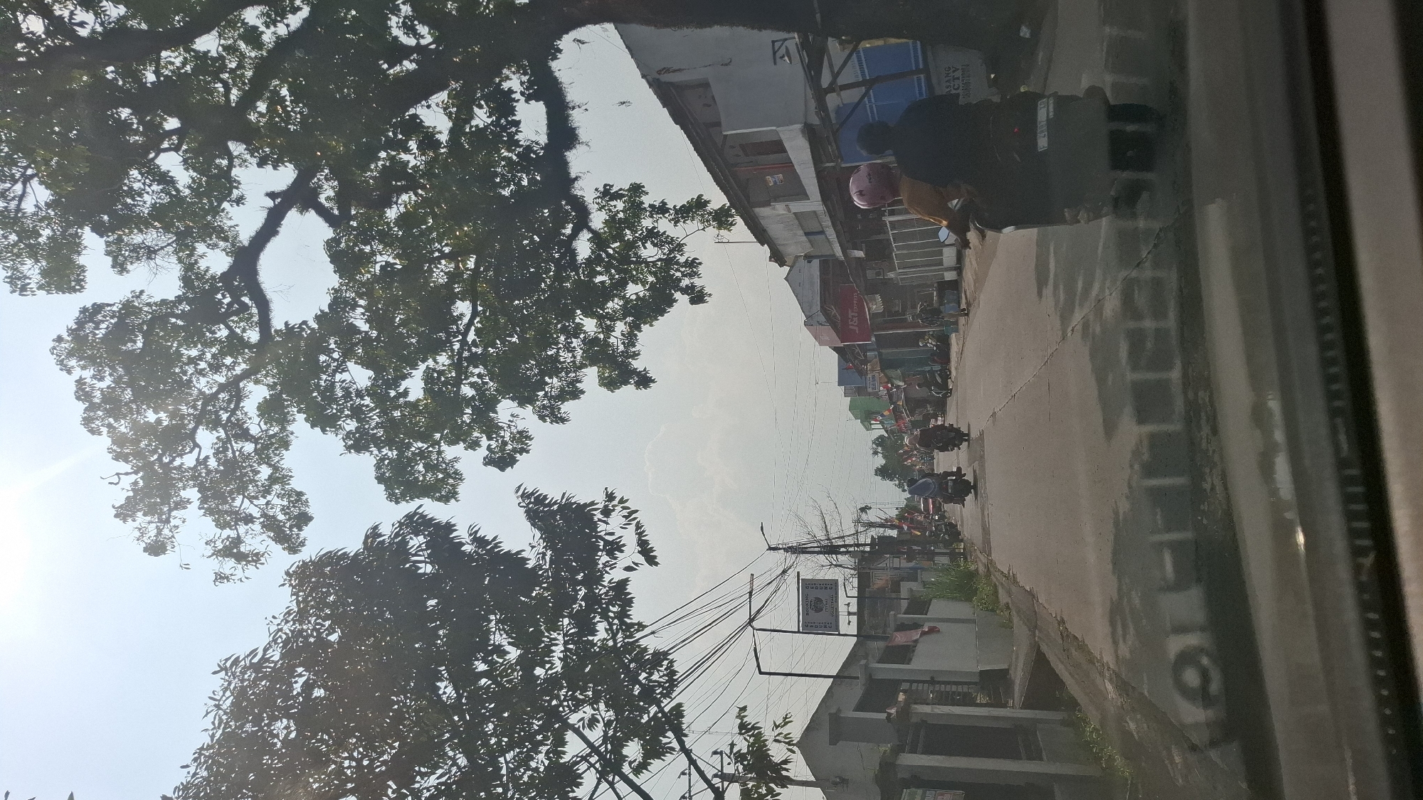 The image depicts a street scene in Cikaroya, Indonesia. The viewpoint is from inside a vehicle, looking forward through the windshield. The road is paved and appears somewhat wet or reflective, with visible cracks. On either side of the road are buildings, some appearing to be shops or homes, with various signs and architectural styles. Vegetation, including large trees with dense foliage, frames the top of the image and encroaches on the sky.
Several people are visible, mostly on motorcycles. Two individuals are riding a scooter in the foreground, with the passenger wearing a pink helmet and the driver wearing a dark top. Further down the road, other motorcycles are also carrying passengers. Some individuals are seen standing or walking along the sidewalks. Electrical wires are strung across the street, supported by poles. The sky is bright and partly cloudy, with the sun's glare visible. One building on the right side of the road has a blue tarp or awning. A sign for "J&T Express" is visible on a building. The overall impression is of a typical day on a moderately busy street in a residential or commercial area of Cikaroya.
