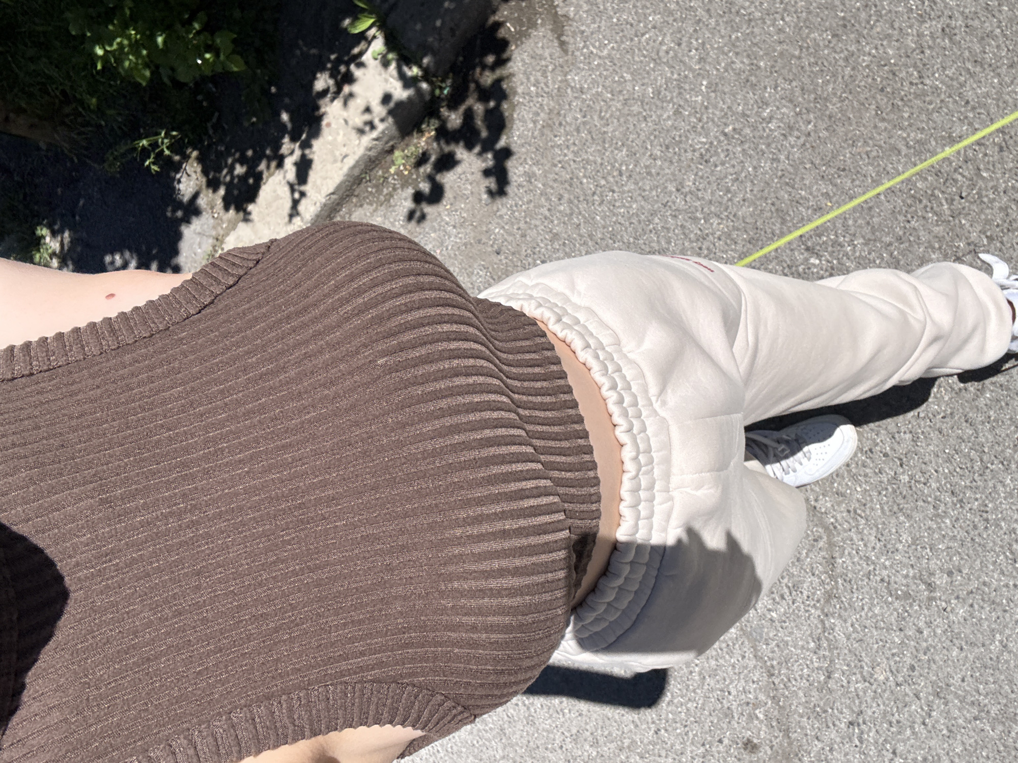 A top-down shot captures a person wearing a brown ribbed tank top and beige sweatpants. Their lower torso and legs are visible against a backdrop of asphalt pavement. To the side, there is foliage, which suggests an outdoor urban setting. The sweatpants are gathered at the ankles. The person is wearing white sneakers. A yellow object is visible in the top right corner of the image. The location in Sofia, Bulgaria, cannot be determined from this image.