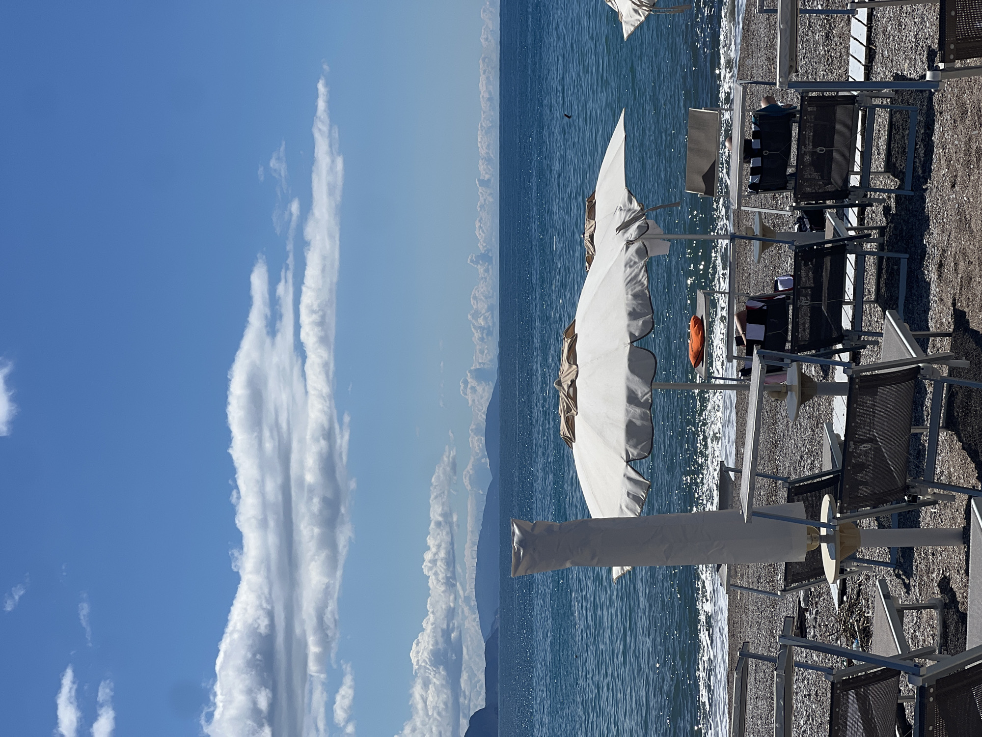 The image shows a beach scene in Prijevor, Montenegro. The beach consists of small pebbles and is lined with rows of beach chairs and partially furled umbrellas. The foreground shows a close view of beach chairs. The background features a calm, blue sea. In the distant background, are clouds along with the faint presence of mountains. A sliver of blue sky with vertical clouds is visible on the far left side of the image. The image is taken from a perspective close to the ground.