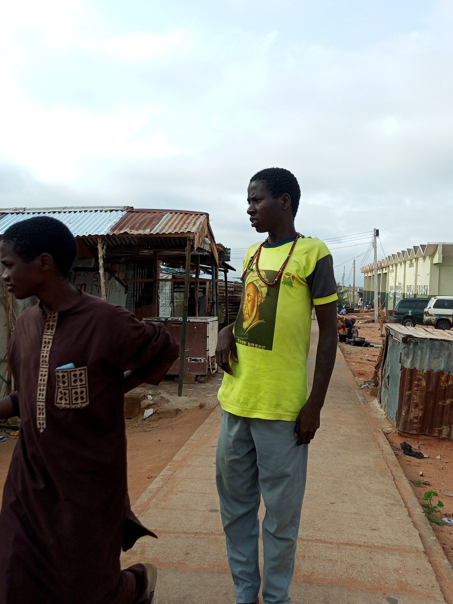 The image captures a street scene in the city of Gombe, Nigeria, under an overcast sky. Two young men are the central figures. The man on the left, in the foreground, is seen from the side, wearing a brown traditional garment adorned with intricate white and tan geometric patterns. He has a dark complexion and short, dark hair, with a visible scar on his forehead. He appears to be walking or standing with his left hand on his hip.
To his right, a second young man faces the camera, his body angled slightly away. He wears a bright lime green t-shirt with dark grey sleeves, featuring a large graphic print of a portrait of a man with a beard and the text "DAVE BEATS" below it. He also wears a necklace of reddish beads. His pants are light grey. He stands with his right hand in his pocket and his left arm bent at the elbow.
The background reveals a street lined with small, makeshift shops with corrugated metal roofs, some appearing weathered and rusty. Beyond these structures, modern buildings with a distinctive tiered roofline are visible, along with parked vehicles, including what appears to be a white SUV and a dark green minivan. The ground is a mix of concrete paving and sandy, reddish earth. In the mid-ground, other individuals and activity are discernible, suggesting a bustling local area. The overall impression is one of a daily life scene in a commercial or residential district within Gombe.