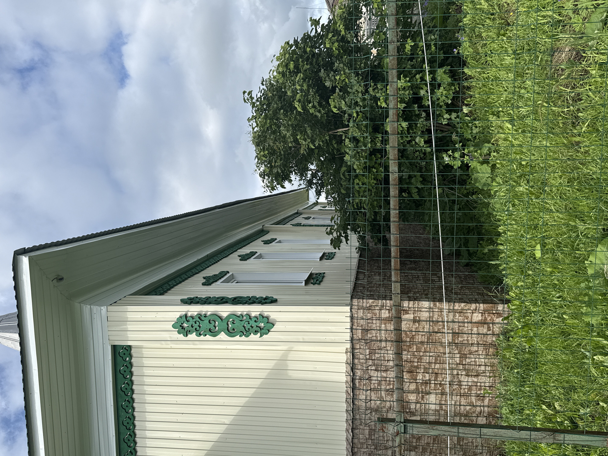 This is a medium shot, taken outdoors, of the side of a light-colored, paneled house with green decorative trim. The house has a white roof with a dark gray ridge. Several windows, also white, are visible on the side of the house. The house appears to be in a rural or suburban setting, as there is a large tree with green leaves and a wire fence with a brick wall behind it. A patch of tall green grass is visible to the right of the fence. The sky is cloudy with patches of blue. The image is oriented vertically. The scene appears to be peaceful and sunny, suggesting it was taken during the daytime. There is no visible text in the image.
