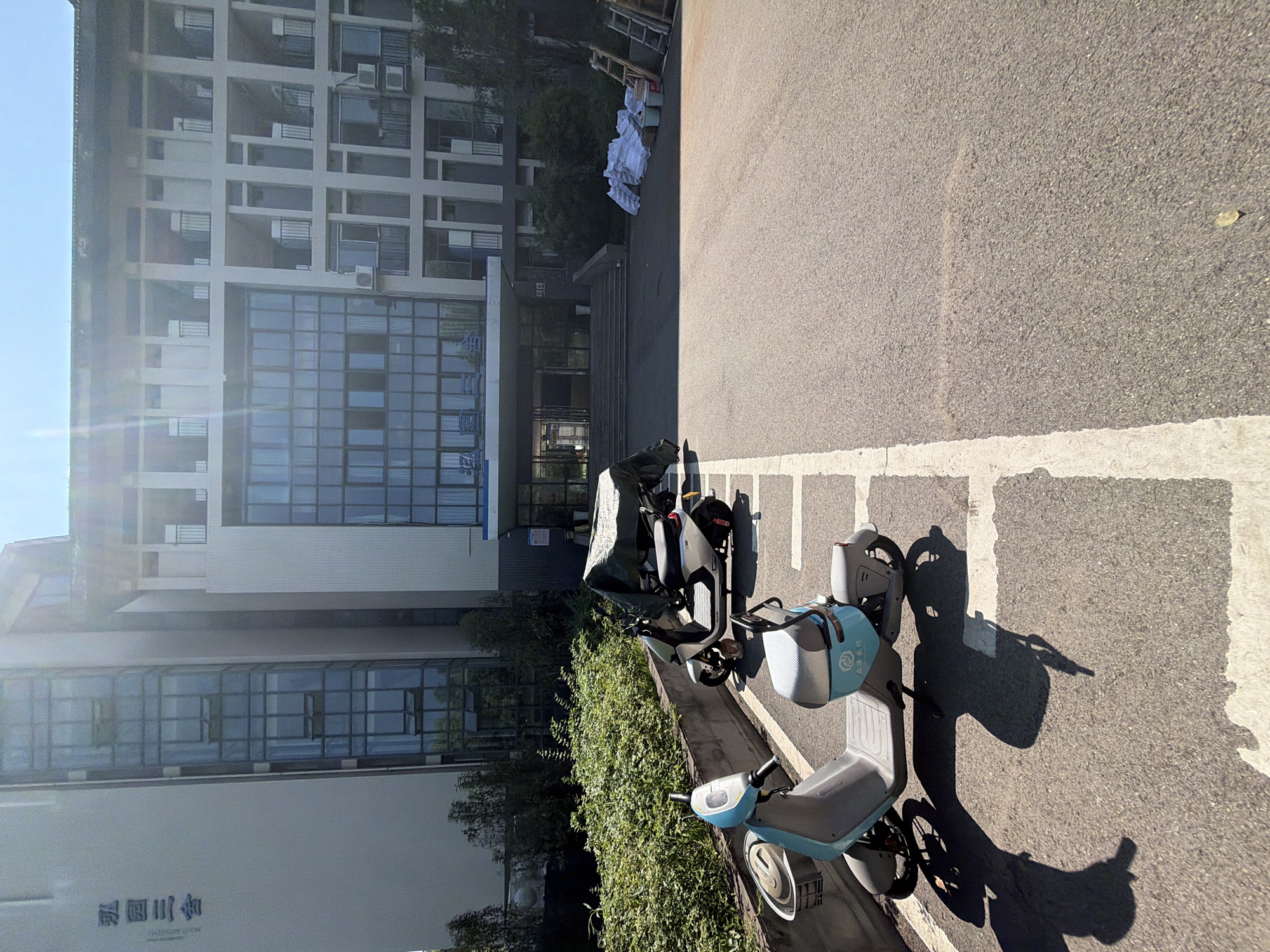 The image shows an outdoor scene in front of a modern building, likely a dormitory, located in the city of Jiangjin, China. In the foreground, two electric scooters are parked. The scooter on the left is light blue and white, with a basket at the front. The scooter on the right is silver and white and is partially covered with a dark tarp. The scooters are parked in designated spots marked by white lines on an asphalt surface. Behind the scooters, there is a low concrete wall and a dense green hedge. The building in the background is multi-story, featuring a modern architectural style with large glass windows and balconies. The entrance to the building has a set of stairs leading up to a covered entryway. To the right of the building entrance, there are piles of white bags, buckets, and ladders, suggesting some sort of construction or maintenance work is underway. The sky is clear and blue, indicating a sunny day. The overall environment appears to be a campus or residential area.