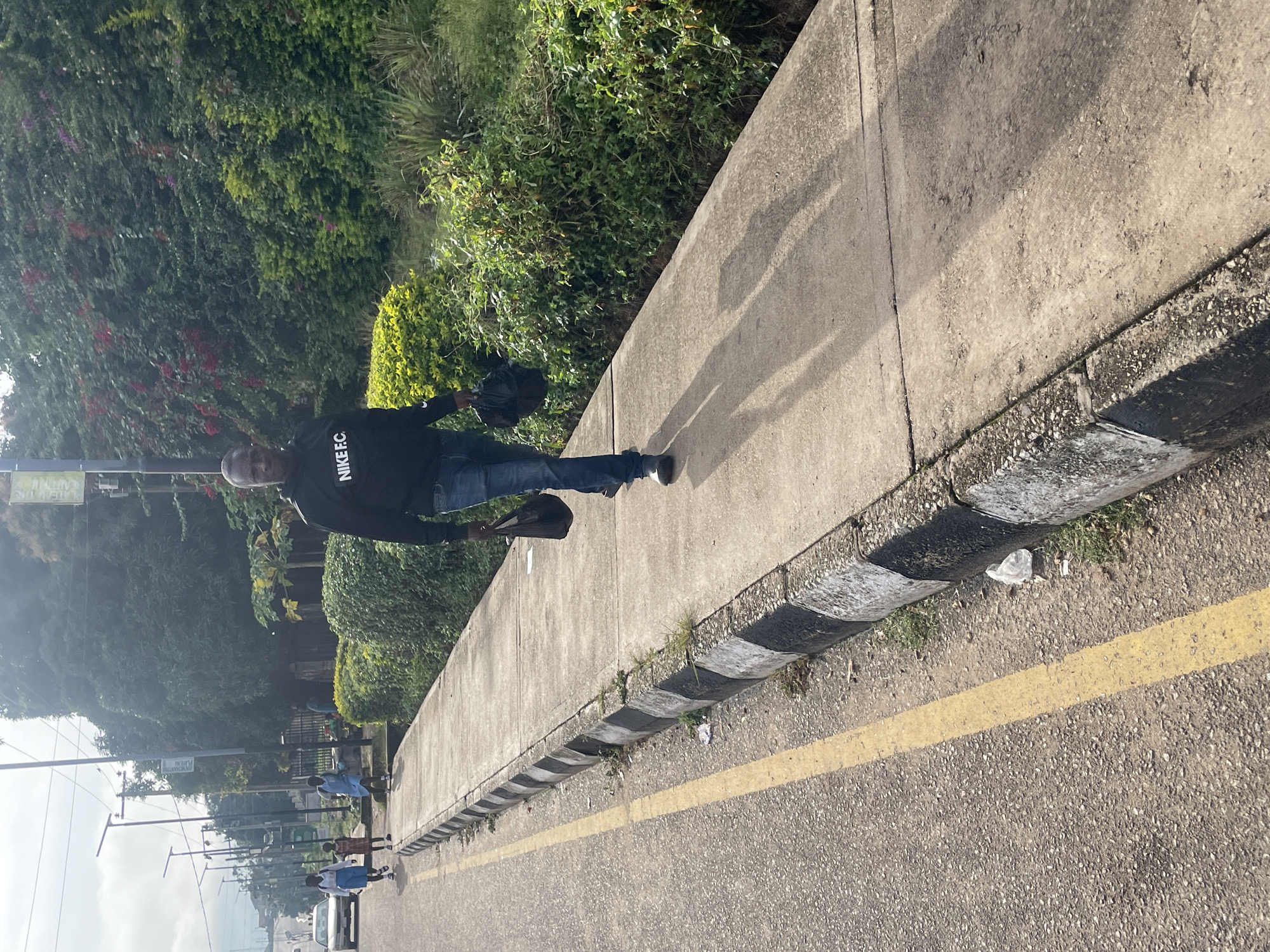 A man walks down a concrete sidewalk next to a road in Jos, Nigeria. He is wearing a black hoodie with "NIKE F.C." written on it in white letters and blue jeans. He is carrying a black plastic bag in his right hand and a black object in his left hand. The sidewalk has a border of black and white painted stones. The road has a yellow line painted down the middle. On the left side of the road, there are streetlights and some vehicles. On the right side of the sidewalk, there are lush green bushes and some flowering plants. The sky is overcast.