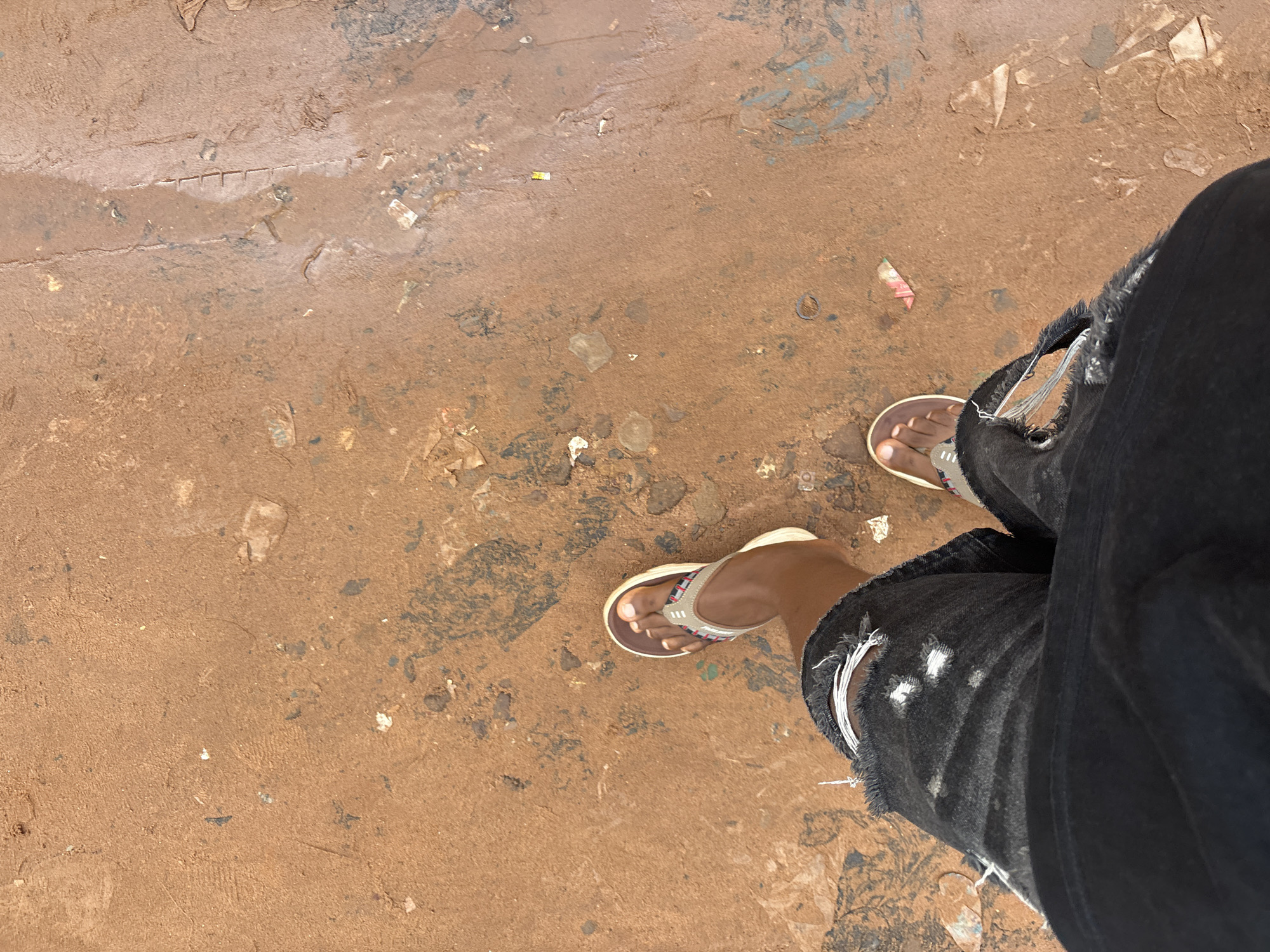 The image shows a person's legs and feet from a high-angle perspective, standing on a dusty, reddish-brown dirt surface littered with small pieces of debris, possibly trash. The person is wearing ripped black denim shorts, with white threads hanging from the rips, and brown and white flip-flops. One flip-flop is visible on the left foot, and the right foot is partially obscured. The ground appears uneven and somewhat muddy in places, with tire tracks or distinct lines visible, suggesting it might be a street or pathway in Onitsha, Nigeria. The overall scene conveys an urban, outdoor environment, likely a public space.