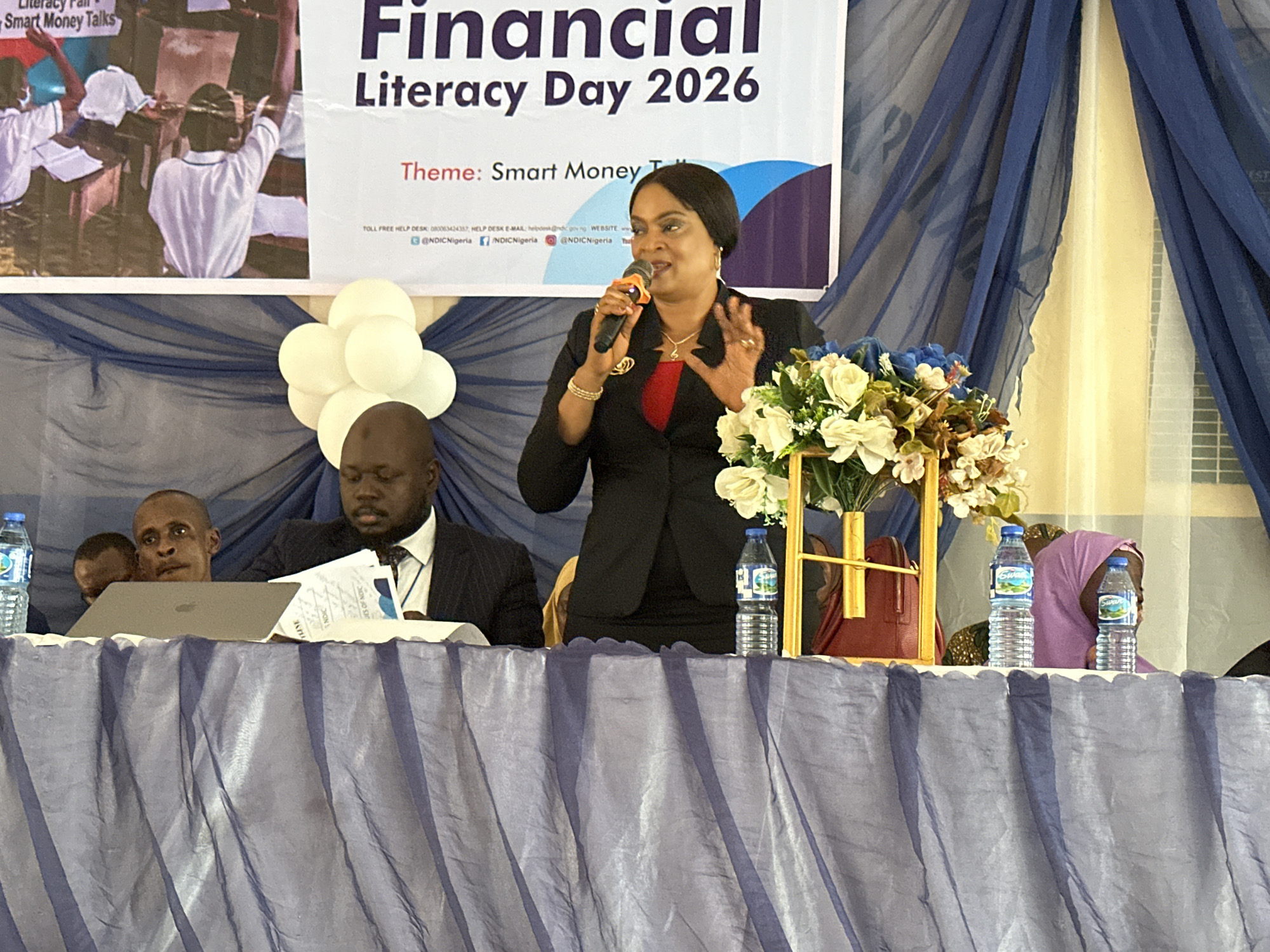 This image depicts an indoor financial literacy event, likely a conference or seminar, held at Unguwan Dosa, Nigeria. The central focus is a woman, presumably a speaker, standing behind a draped table and addressing an unseen audience. She is dressed in a black blazer over a red top, with a dark head covering, holding a microphone in her right hand and gesturing expressively with her left. Her hands are adorned with rings and bracelets.
Seated to her left at the table is a man in a dark suit and tie, looking down at documents and a laptop. Another man's head is partially visible behind him. On the far right, a woman in a purple hijab is partially visible. The table is covered with a pleated, translucent purple-blue fabric. Objects on the table include a laptop, various papers, two bottles of "Swan" brand water, and a floral arrangement featuring white and blue artificial flowers. A cluster of white balloons can be seen to the left of the speaker.
In the background, a large banner announces "Financial Literacy Day 2026" with the theme "Smart Money Talks." The banner also displays contact information: "TOLL FREE HELP DESK: 080063424357," "HELP DESK E-MAIL: helpdesk@ndic.gov.ng," and social media handles such as "@NDICNigeria." An image of children in a classroom, with the text "Smart Money Talks," is also part of the banner's design. The venue's background is decorated with blue and light-colored sheer drapes, suggesting a formal or semi-formal setting. The event appears to be organized by or in collaboration with "NDIC."