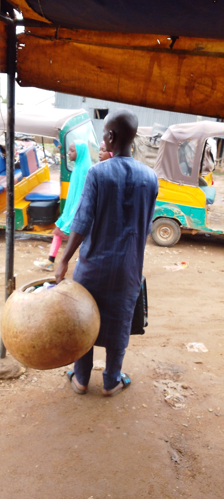 A medium-shot image depicts a person walking away from the viewer, carrying a large gourd. The individual is bald, wearing a dark blue striped tunic and pants, and blue sandals. The gourd, held in the right hand, appears to be full of items. Behind the person, two girls stand near yellow and green auto rickshaws. One girl wears a light blue hijab. The ground is dirt, with some litter visible. The scene is partially shaded by an overhang. The location is presumed to be in Jos, Nigeria, based on the cultural clothing and type of transportation.