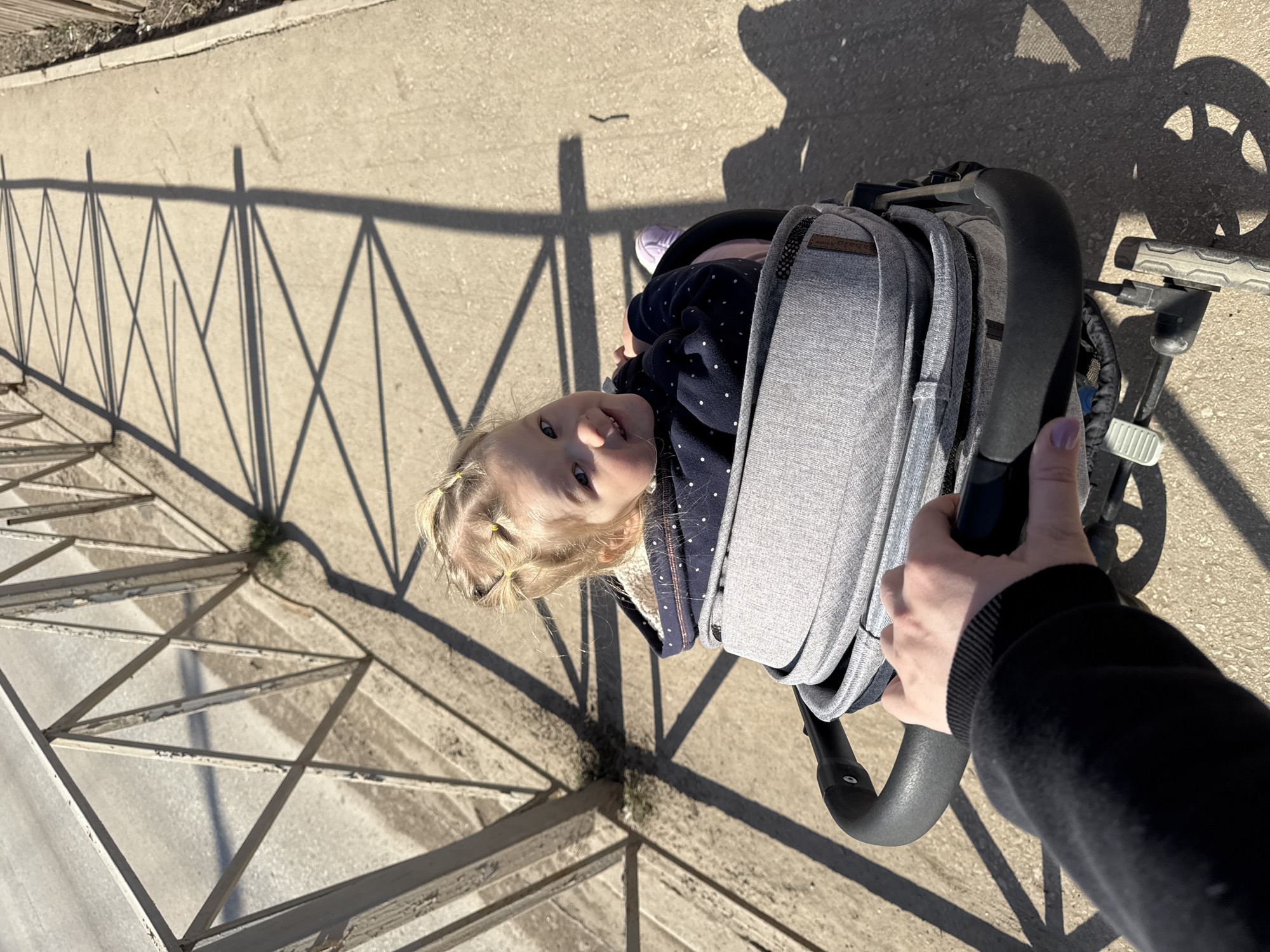 The image captures a high-angle view looking down at a young child in a grey stroller, being pushed outdoors on a sunny day in Ферма, Russia. The perspective suggests the photograph was taken by the person pushing the stroller, whose right hand is visible on the stroller's black handle in the lower right corner.
The main subject is a fair-haired child, likely a toddler, with blonde hair styled in two small pigtails secured with yellow hair bands. The child wears a navy blue long-sleeved top with white polka dots and looks directly up at the camera with a curious, slightly smiling expression. A purple shoe is visible on the child's foot. The stroller itself is made of grey fabric with a dark frame, and a small tan label, possibly reading "BabaBoom," is visible on its side near the child's head.
The setting is an outdoor paved area, likely a sidewalk or path, alongside a metal fence. The fence, featuring a repeating crisscross pattern, casts long, sharp shadows across the light-colored pavement, indicating strong sunlight, consistent with either morning or late afternoon. These shadows create a prominent geometric design on the ground. Another shadow, likely from the stroller's wheel, is visible in the upper right. The scene conveys a bright, clear day.