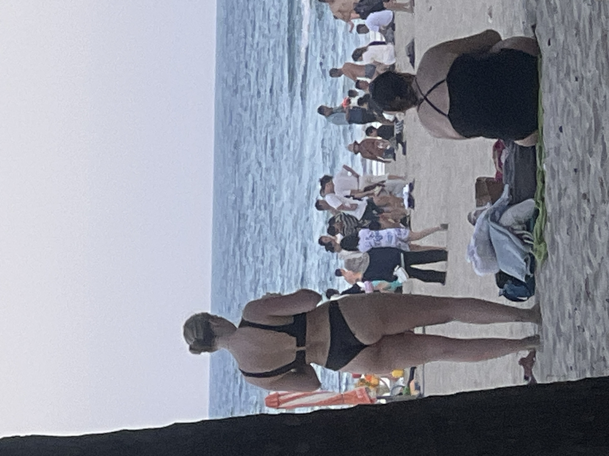 This media file shows a crowded beach scene in Da Nang, Vietnam, likely during the day with overcast or hazy skies. The primary subjects are numerous people, many of whom are wearing swimwear. The scene is dominated by the sandy beach and the ocean, with waves visible.
In the foreground, a woman is captured from behind, wearing a black bikini. She is standing on the sand. Further back, another woman in a black swimsuit is sitting on the sand, also facing away from the camera.
The middle ground and background reveal a multitude of people engaged in various beach activities. Some are standing, some are sitting, and some appear to be walking or interacting. The clothing suggests casual beach attire, including swimwear, t-shirts, and shorts. The presence of beach towels and some folded clothing indicates people relaxing or preparing to swim.
The general atmosphere appears to be lively and social, typical of a popular beach destination. No specific time of day can be precisely determined beyond it being daytime. The weather seems to be a bit hazy or overcast, as the sky is a pale, uniform color, and there are no harsh shadows. The overall impression is one of a busy, communal beach experience in a tropical or subtropical location. There is no visible text in the image.
