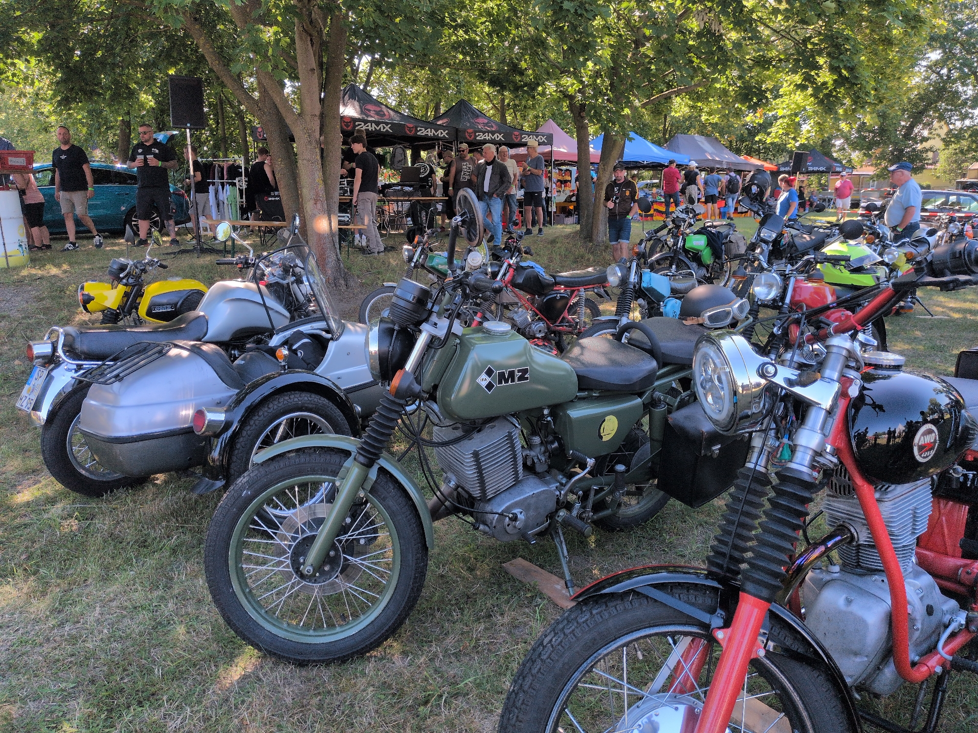 The image captures an outdoor gathering, likely a motorcycle event, on a sunny day. In the foreground, several vintage motorcycles are parked on a grassy area beneath trees. One prominent motorcycle in the center is olive green with the "MZ" logo visible on its fuel tank. To its left, another motorcycle with a silver sidecar is parked. The background reveals a row of tents, with "24MX" branding visible on some. Various individuals, dressed casually, are seen milling around the tents and motorcycles. Some are standing, talking, while others appear to be browsing. A blue car is parked to the left of the frame. The overall atmosphere suggests a community event focused on motorcycles, set in a park-like environment. The location is identified as Hohenferchesar, Germany.
