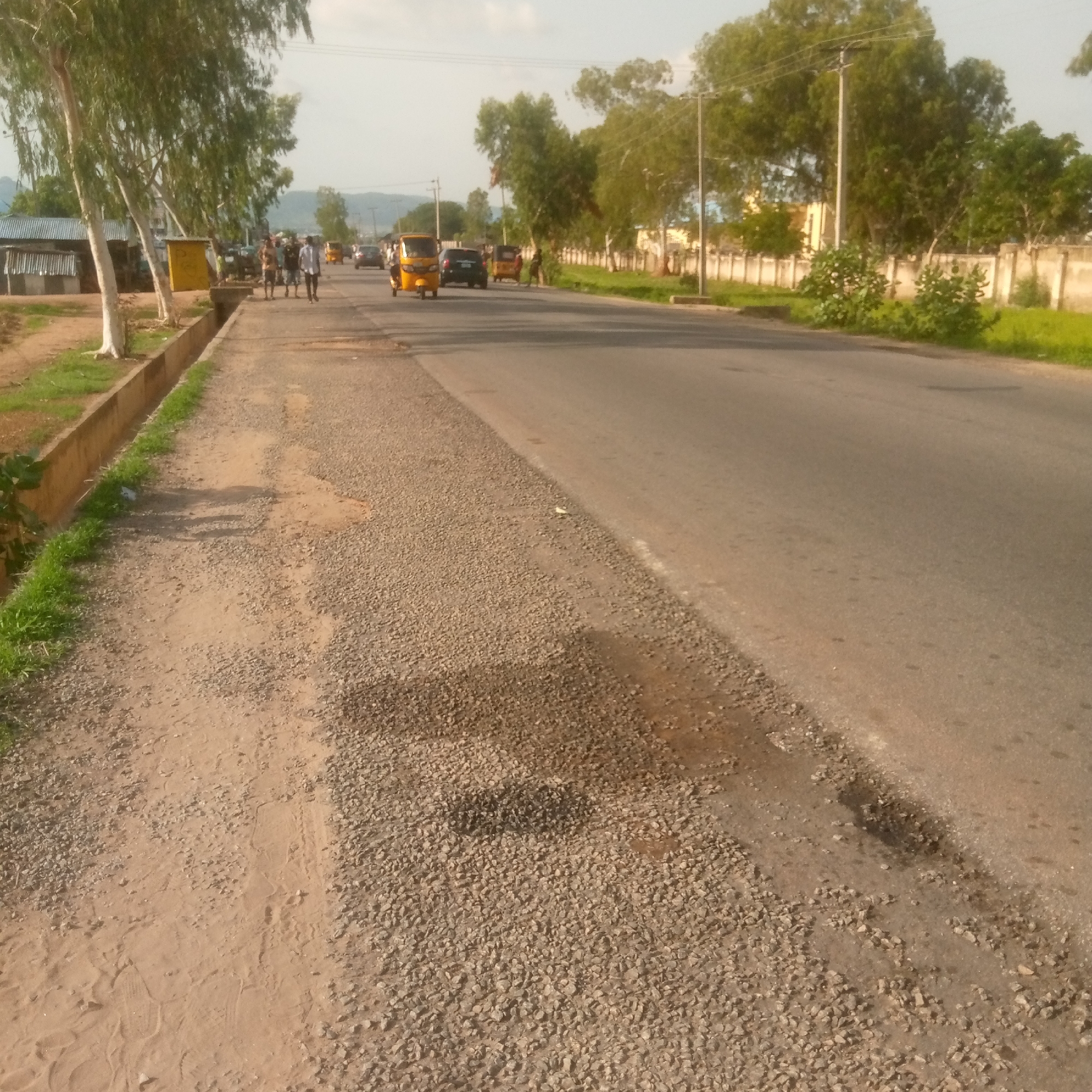 A street scene with several vehicles and people is captured. A yellow auto-rickshaw is in motion on the asphalt road, accompanied by other vehicles and pedestrians walking along the side of the road. The road has a rough gravel shoulder and a concrete drainage ditch runs alongside it, with some grass growing in and around it. Trees line the opposite side of the road, with a wall and buildings further in the background. The time of day appears to be late afternoon or early evening, as indicated by the warm, low-angled sunlight casting shadows. The sky is partly cloudy. The overall mood is one of everyday activity in a semi-urban or rural setting. There are no visible signs or text in the image.