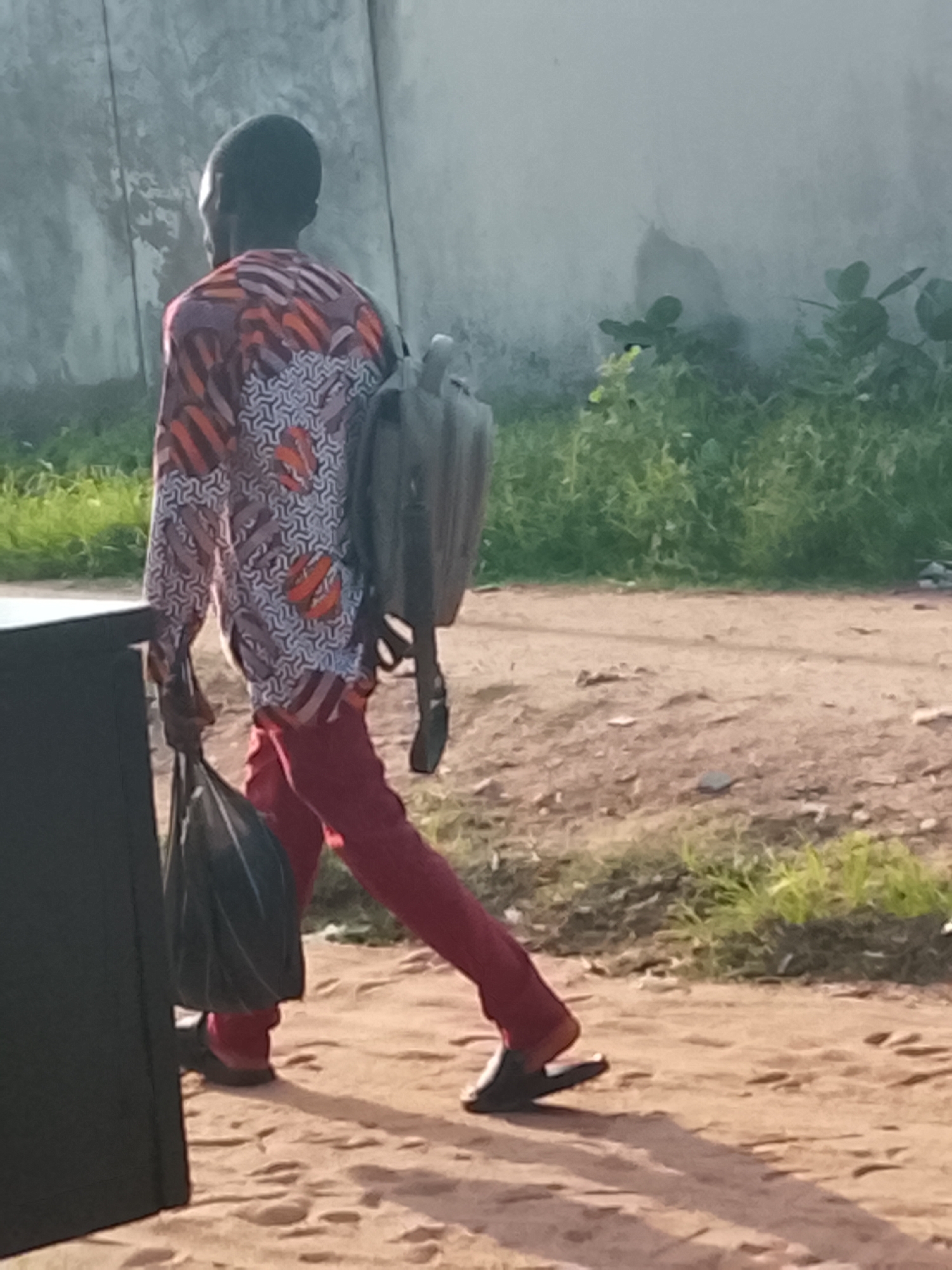 A Black male, appearing to be in his late 20s or early 30s, is walking away from the camera. He is wearing a long-sleeved shirt with a colorful, patterned design in shades of purple, orange, and white. He is also wearing red pants and dark slip-on shoes. On his back, he carries a large, rectangular backpack. In his left hand, he is holding a black plastic bag. The man is walking on a dirt path, which appears to be in an outdoor setting with sparse vegetation and a concrete wall in the background. The lighting suggests it is daytime, possibly late afternoon due to the shadows. The environment appears to be a rural or semi-urban area within Jalingo, Nigeria.