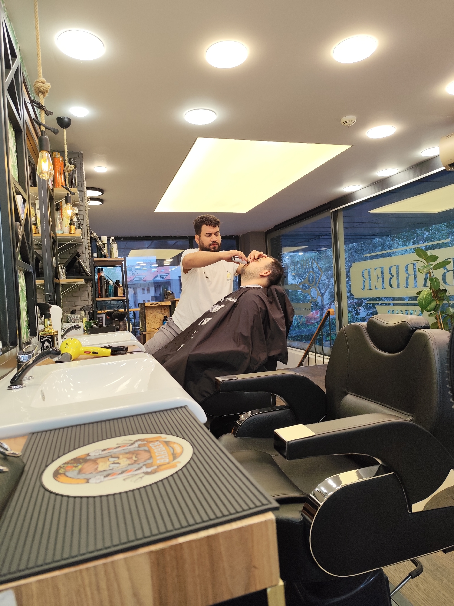 A barber is trimming the beard of a client in a well-lit barbershop. The barber, wearing a white t-shirt, is holding clippers to the client's face. The client is seated in a black barber chair, covered with a black cape. The barbershop has modern lighting, including circular ceiling lights and a large illuminated panel. To the left, a white sink with a chrome faucet and a yellow hairdryer are visible. Shelves with various bottles and products line the back wall. A large window in the background shows a glimpse of the outdoors with trees and buildings, suggesting the location is in Sarıyer, Turkey. The overall atmosphere is clean and professional, with a focus on customer service and grooming.