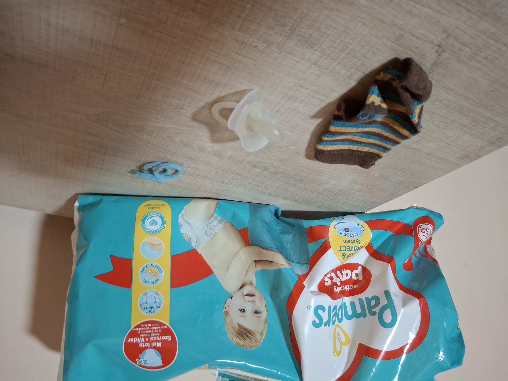 An overhead shot shows a wooden surface with a package of Pampers diapers and baby items. The Pampers package, which is teal, red, and white, is to the left and slightly angled. It features the Pampers logo, an image of a baby, and text in multiple languages, including "active baby pants" and "2x Mai late Szersze Wider". To the left of the package is a single, brown and striped sock with yellow and blue accents, lying on its side. In the center of the frame, a white pacifier rests on the surface. To the right of the pacifier is a small, looped piece of light blue yarn. The scene appears to be indoors, likely on a table or counter, as indicated by the wooden surface and the background wall. No people are present in the image.