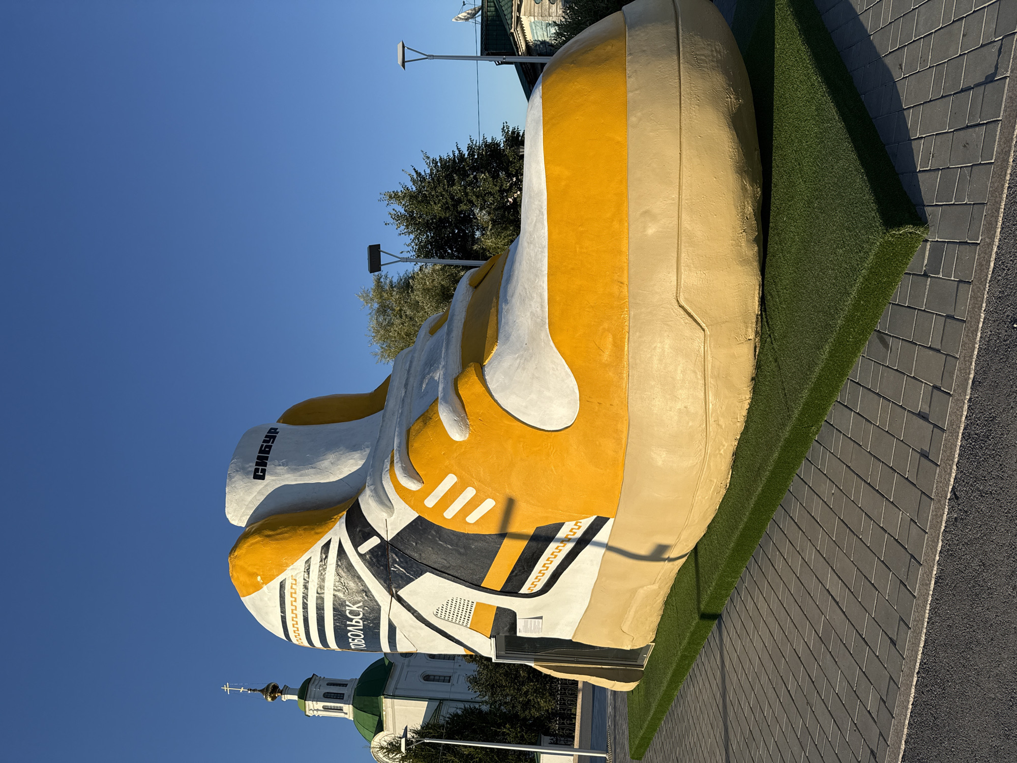 A giant, yellow and white sneaker sculpture stands on a cobblestone plaza in Tobolsk, Russia. The sculpture has "СИБУР" and "ТОБОЛЬСК" written on it. Behind the sculpture, a white church with a green dome and a cross on top is visible. The sky is clear and blue. There are trees and streetlights in the background. The ground is paved with grey rectangular stones, and a section of artificial grass is placed around the base of the sculpture. The sculpture is illuminated by the sun, casting a shadow to the right. There are no people visible in the image.