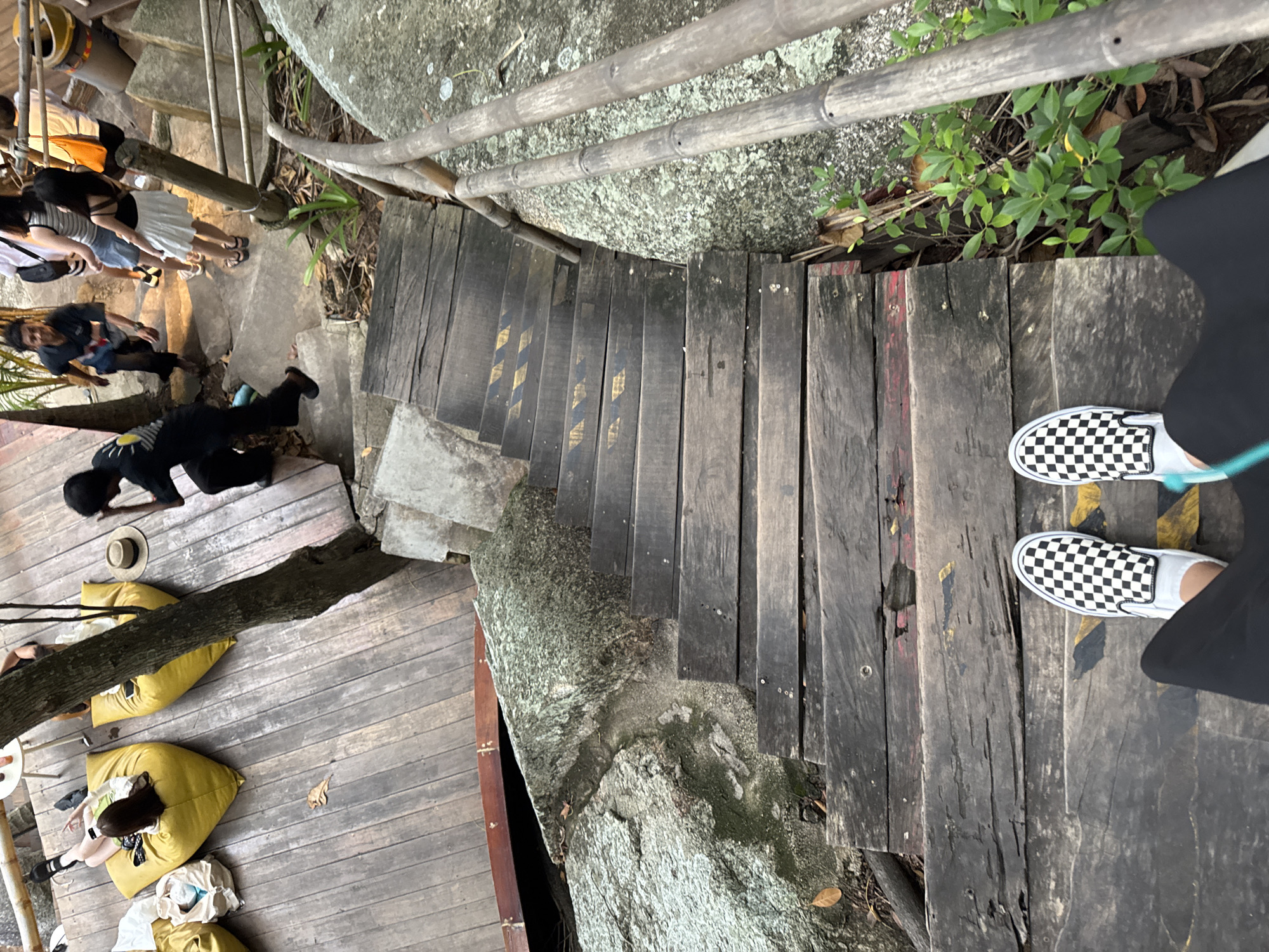 This image shows a wooden staircase and walkway in a natural outdoor setting. Several people are visible, some walking down the stairs and others sitting or lying on yellow beanbag chairs on the wooden platform below. A person wearing black pants and checkered slip-on shoes is standing on the stairs, looking down. The stairs are made of weathered dark wood, with some yellow and black hazard markings on them. The walkway appears to be built around large rocks and trees. Green foliage is visible in the background, suggesting a tropical environment. The overall impression is of a relaxed, casual gathering spot, possibly a scenic viewpoint or a resting area in a park or resort. The lighting suggests it is daytime, but the exact time is difficult to determine. There is no visible text in the image. The location context of Karon, Thailand, further supports the assumption of a warm, possibly humid, outdoor environment.