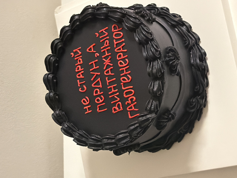 A round cake is positioned centrally on a white, flat display surface. The cake is uniformly frosted in black, featuring a smooth matte finish on its top and sides. Intricate decorative borders of dark piped frosting, consisting of swirls or rosettes, adorn both the top edge and the base circumference of the cake. Piped in bright red frosting on the cake's top surface is the Cyrillic text "не старый пердун, а винтажный газогенератор," which translates to "not an old fart, but a vintage gas generator." The background consists of a plain, light-colored wall, likely off-white or beige, indicating an indoor setting. No individuals or other objects are present in the visible frame. The scene is located in Tel-Aviv, Israel.