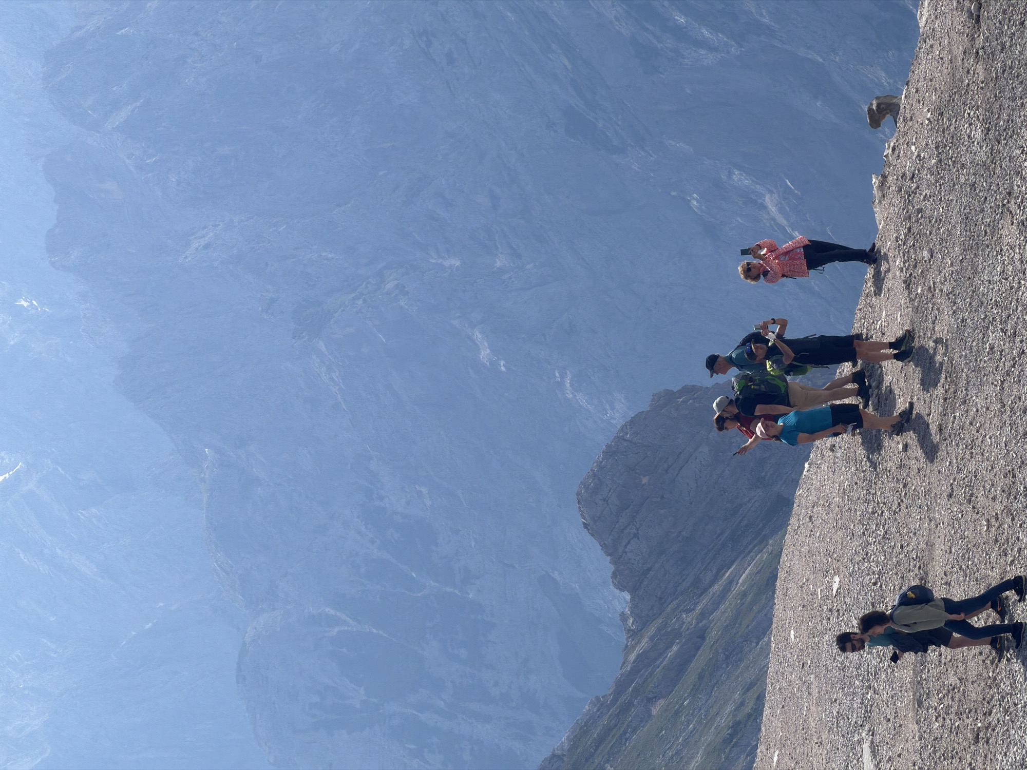 A group of tourists are hiking in the mountains near Garmisch-Partenkirchen, Germany. The image shows a steep, rocky terrain with a panoramic view of hazy blue mountains in the background. On the right side of the image, a woman in a pink and white patterned jacket is taking a picture with her phone. Next to her, a man in a blue shirt and dark shorts is also taking a picture with his phone. Further left, a group of three hikers are walking away from the camera. One man in a blue shirt and dark shorts has a backpack on. The person next to him, wearing a green long-sleeved shirt, also has a backpack. The third hiker is a woman in dark pants and a light-colored shirt, also with a backpack. In the foreground, two hikers are walking towards the camera; one in a dark shirt and shorts, the other in a light-colored long-sleeved shirt and dark pants. The weather appears to be clear and sunny, with a slight haze obscuring the distant peaks. The overall impression is of a scenic mountain hike.
