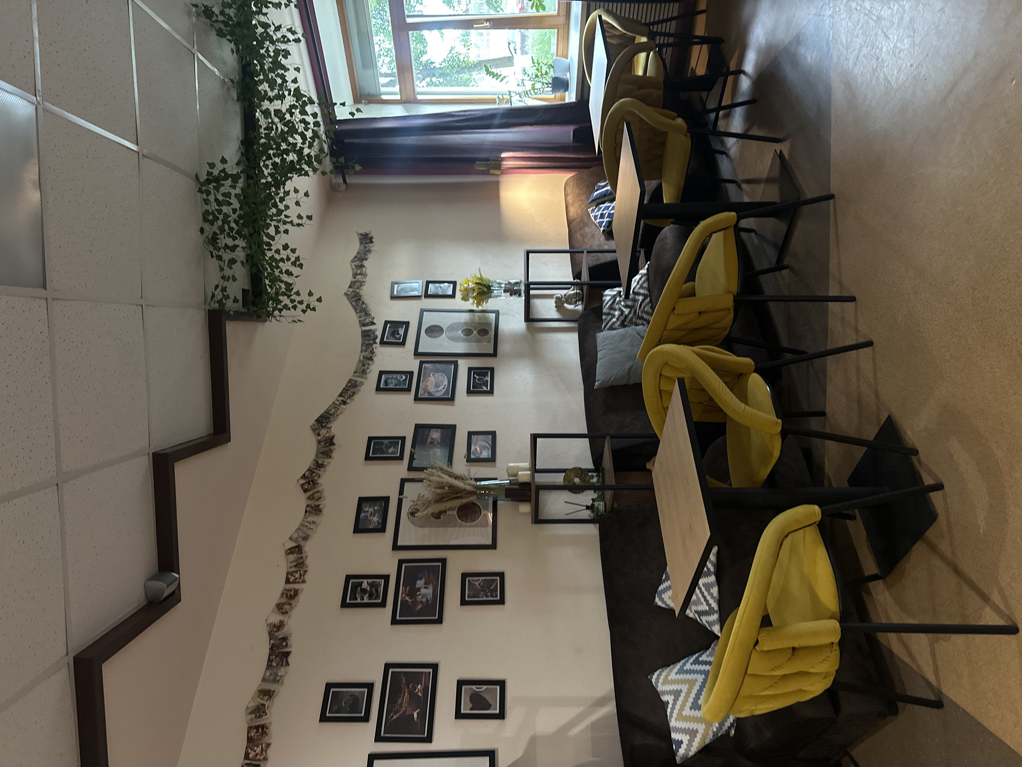 The image depicts the interior of a cafe in Votkinsk, Russia. The cafe features a warm and inviting atmosphere with light-colored walls adorned with numerous framed pictures and a decorative garland of small photos. A dark brown sofa runs along the wall, accented with patterned throw pillows. Several yellow upholstered chairs surround dark-colored square tables with light wood tabletops.
The room is lit by a combination of ambient light, possibly from the window on the right, and artificial lighting. Green artificial ivy vines are draped along the ceiling, adding a touch of nature to the space. A tall window on the right side of the image reveals a glimpse of the outdoors, showing greenery and trees, suggesting the cafe is in an urban or suburban setting.
On a shelf unit against the wall, there is a vase with dried flowers, candles, and a diffuser. The floor appears to be a textured, light brown material. The overall impression is that of a cozy and well-decorated cafe, possibly a popular spot for relaxation and socializing.