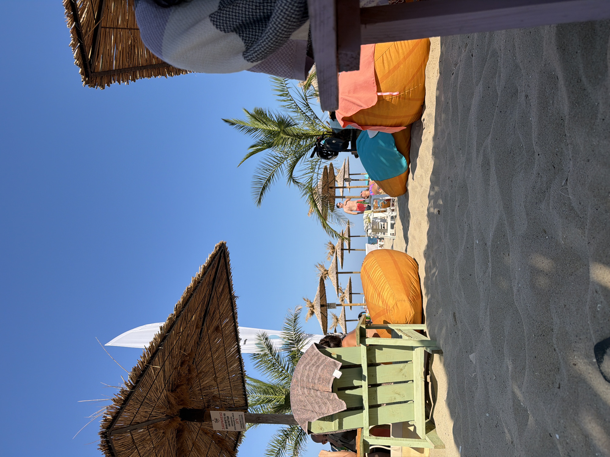 The image depicts a sunny beach scene on a clear day, likely in Varna, Bulgaria, with an emphasis on leisure and relaxation. The foreground is dominated by sand, with shadows cast by unseen objects. In the mid-ground, several elements suggest a resort or public beach setup.
To the left, a green wooden chair is positioned under a straw umbrella, with a striped towel draped over its back. A sign, partially visible on the umbrella's pole, contains Cyrillic text, suggesting a location where Bulgarian is spoken. Beyond this, large, orange beanbag chairs are scattered on the sand, some covered with brightly colored fabrics (blue and pink). Palm trees are visible behind these seating arrangements, indicating a warm climate.
In the background, more straw umbrellas and lounge chairs are visible, stretching towards the sea. A few individuals are present on the beach; one man in red swim trunks stands near the palm trees, while another person is partially visible lying down. Further in the distance, the clear blue sky meets the horizon, hinting at the sea. The overall atmosphere is one of a typical beach day, with amenities for visitors to enjoy the sun and sand.