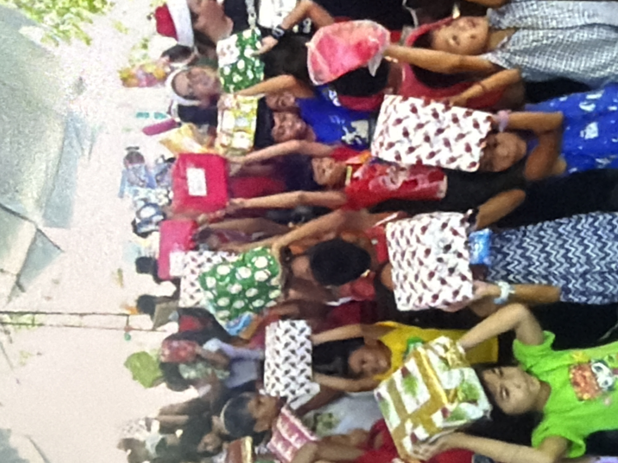 A group of children, most of whom appear to be under the age of ten, are gathered together outdoors, holding wrapped presents above their heads. Many of the children are wearing festive red and white Santa hats, suggesting the event may be related to a holiday celebration or gift-giving occasion. The children are holding a variety of brightly colored and patterned gift boxes. Some of the children are smiling and looking towards the camera, while others appear to be looking at their presents. The background is slightly out of focus but appears to show some trees and possibly a tent or shelter. The lighting suggests it is daytime.
