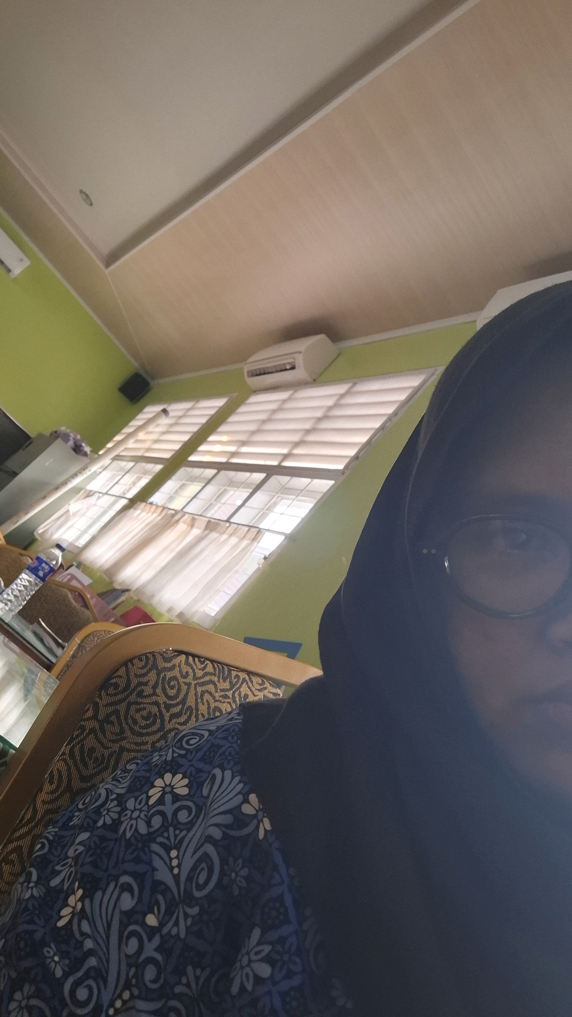 A person wearing a dark hijab and glasses is seated in a room, with the camera positioned to capture their face and upper body. The room has light green walls and a light-colored, slanted ceiling with a visible air conditioning unit mounted on the wall above a set of windows. The windows are covered with light-colored blinds and sheer curtains, allowing diffused light to enter. The person is seated in a chair with a patterned fabric on its backrest and a blue and white floral patterned garment visible on their shoulder. On a table in the background, a bottle of water is present. The image is taken from a low angle, looking upwards towards the ceiling.