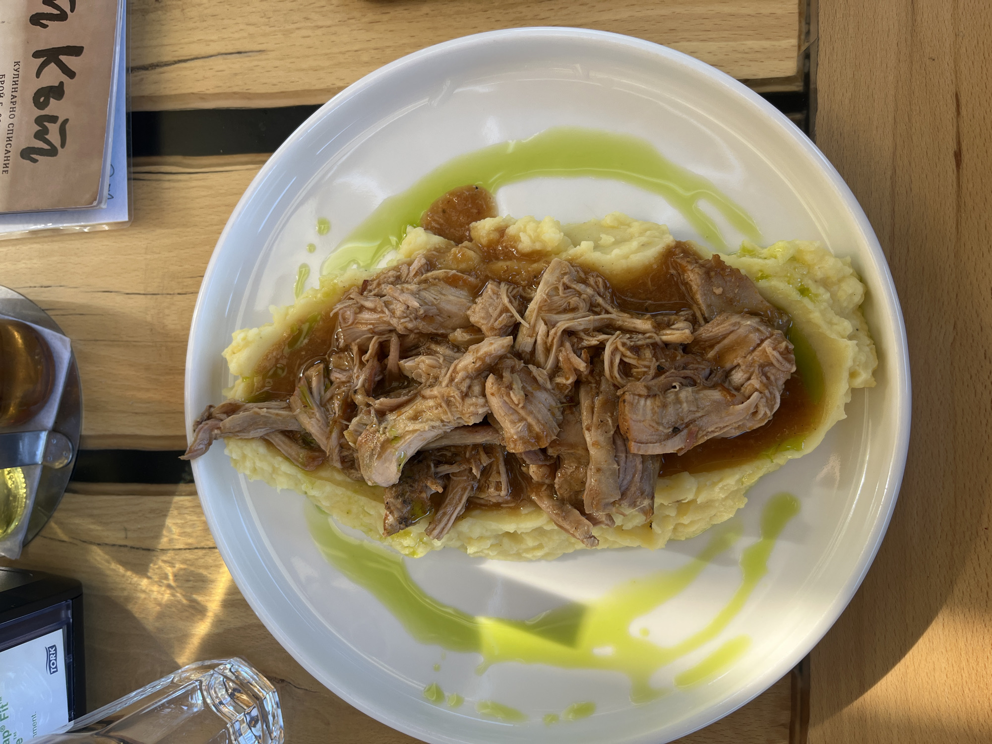 A top-down shot reveals a plate of food on a wooden surface. The plate is round and white, containing a bed of mashed potatoes topped with shredded meat and a sauce. A green drizzle garnishes the plate. Parts of a paper menu, glass, and a black object are visible on the table. The image was taken in Slokoshtitsa, Bulgaria.