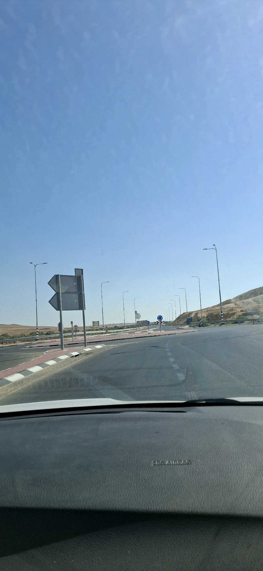 The image depicts a view from inside a vehicle, looking out at a highway intersection in Yeruham, Israel. The sky is clear and blue. The road is asphalt with white dashed lines. There are several streetlights lining the road, and a large directional signpost is visible on the left. The landscape surrounding the road appears arid, with low scrub vegetation and hills in the distance. The foreground shows the dashboard of the vehicle, with the text "SRS AIRBAG" visible.