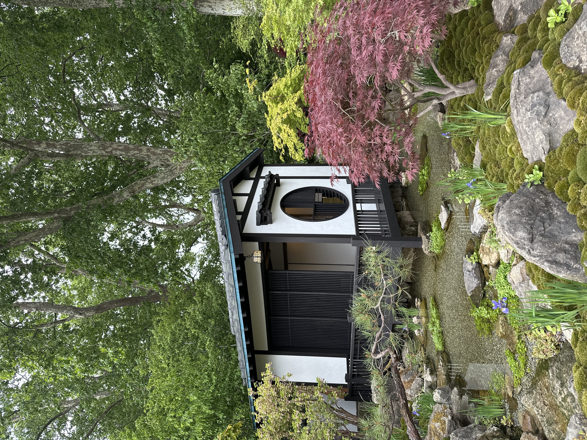 The image depicts a traditional Japanese building, likely a tea house, nestled within a lush garden setting. The building is characterized by its white walls contrasted with dark black trim. It features a distinctive circular window on the upper level and a horizontally slatted design on the lower level. The garden is rich with vegetation, including moss-covered rocks, a variety of trees with green and red foliage, and flowering plants. A small stream or gravel pathway runs alongside the building. The image does not contain any visible people. The overall scene evokes a sense of tranquility and harmony with nature. There is no evidence that the scene is in London, United Kingdom.