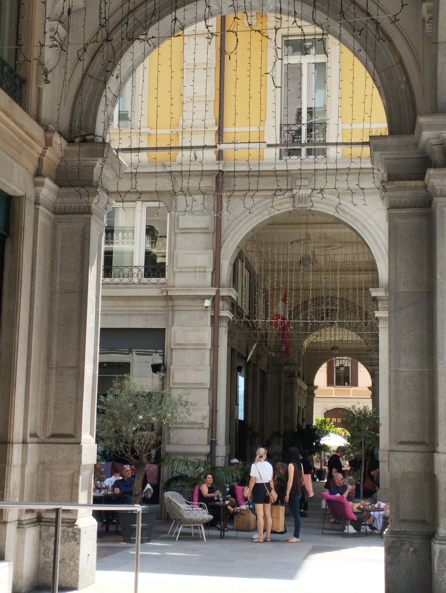 This is a medium shot of an outdoor cafe setting in Lugano, Switzerland, viewed from beneath an arched walkway adorned with strings of lights. In the foreground, stone architectural elements frame the scene. A few people are seated at tables. On the left, a man in a blue shirt sits at a table with other patrons, and an olive tree in a large pot is visible. To the right, a woman in a black dress sits at a table with a pink cushion on a chair. Further back, a woman in a white shirt and black shorts stands, and another woman in a dark top and jeans is barefoot. A man in a blue shirt and pink shorts sits in a pink chair, and other people are seated at tables in the background. The cafe area has a white umbrella and palm plants, with a building facade visible behind it. The overall impression is a sunny, leisurely afternoon in a European city.