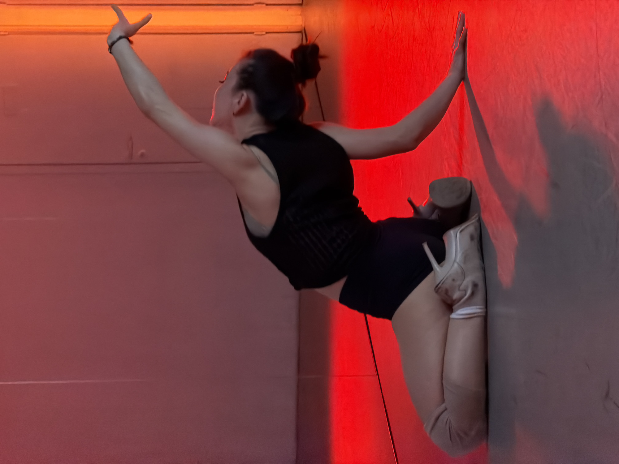 A person wearing a black top and shorts with high-heeled shoes is posing against a wall. The person's hair is in a bun. The wall is lit with red light on one side, creating a shadow effect. The exact location in Moscow, Russia, is not identifiable from the image.