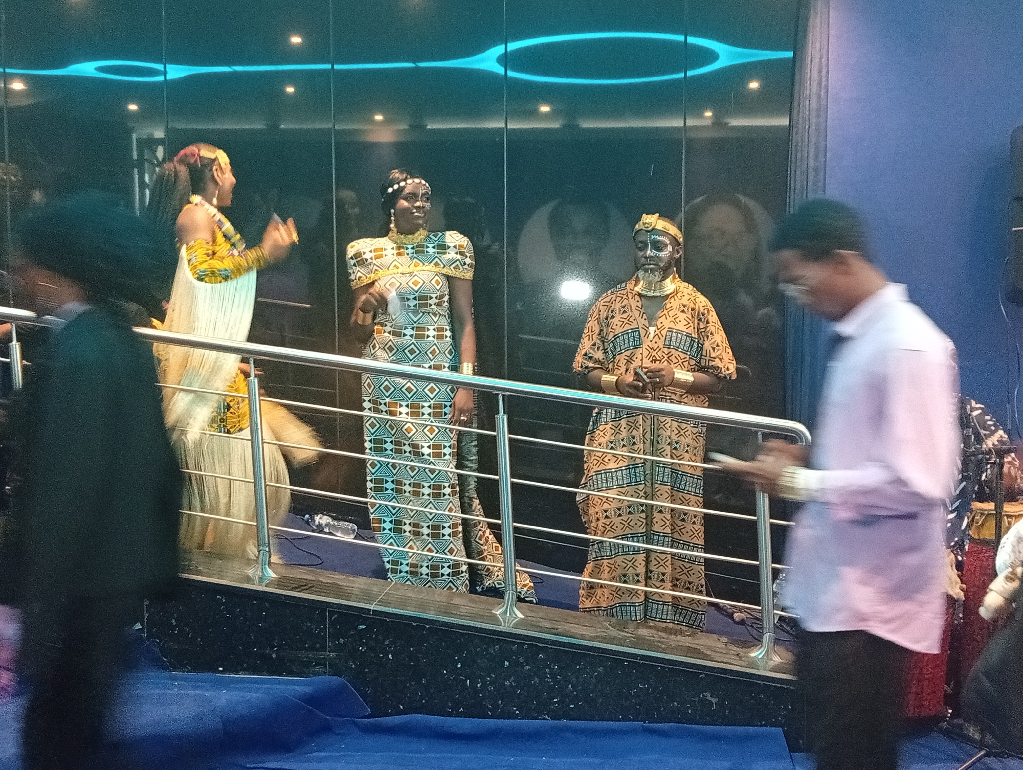 The image shows three individuals in cultural attire standing behind a metal railing. The environment is indoors, possibly a stage or elevated platform, with a blue carpet on the floor. To the right and left are partially visible individuals in modern clothing.
The three people behind the railing are wearing tribal clothing, including beaded necklaces, headdresses, and ornate patterns on their dresses. One individual has painted facial markings. They appear to be presenting or performing. Behind them is a dark, reflective surface, potentially a window.
The lighting is diffused, with some blue highlights.
The location is Itirin, Nigeria.