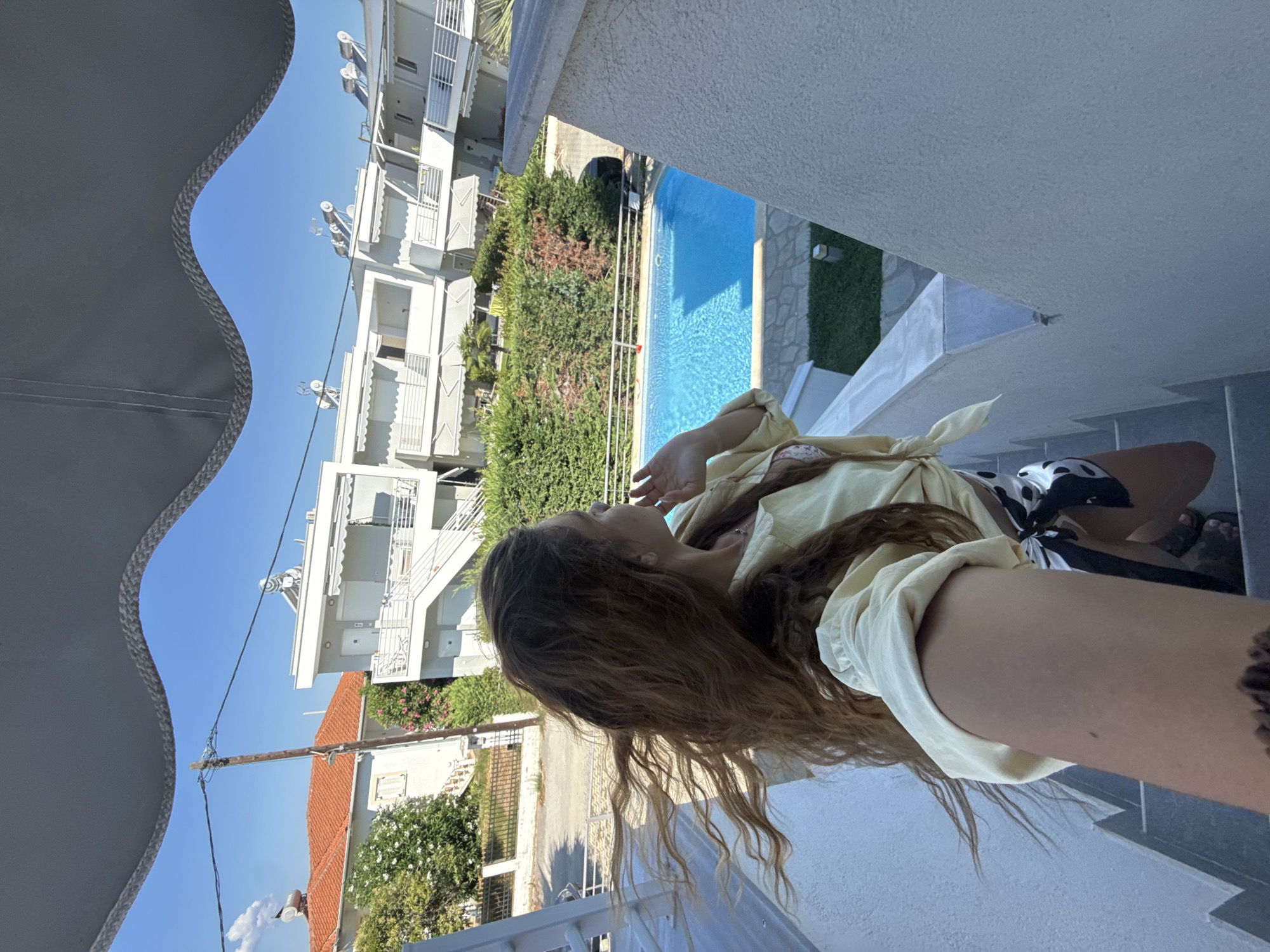 The image shows a young woman standing on a white staircase outside a building with a blue swimming pool and other buildings in the background. The woman is facing away from the camera, looking to her right. She has long, wavy brown hair that is blowing in the wind. She is wearing a yellow shirt tied at the waist and black and white patterned shorts. The staircase is outdoors, with a white railing. The swimming pool is rectangular and filled with bright blue water. In the background, there are several white buildings with balconies and awnings, as well as green bushes and trees. The sky is clear and blue. The scene appears to be in a residential or tourist area.