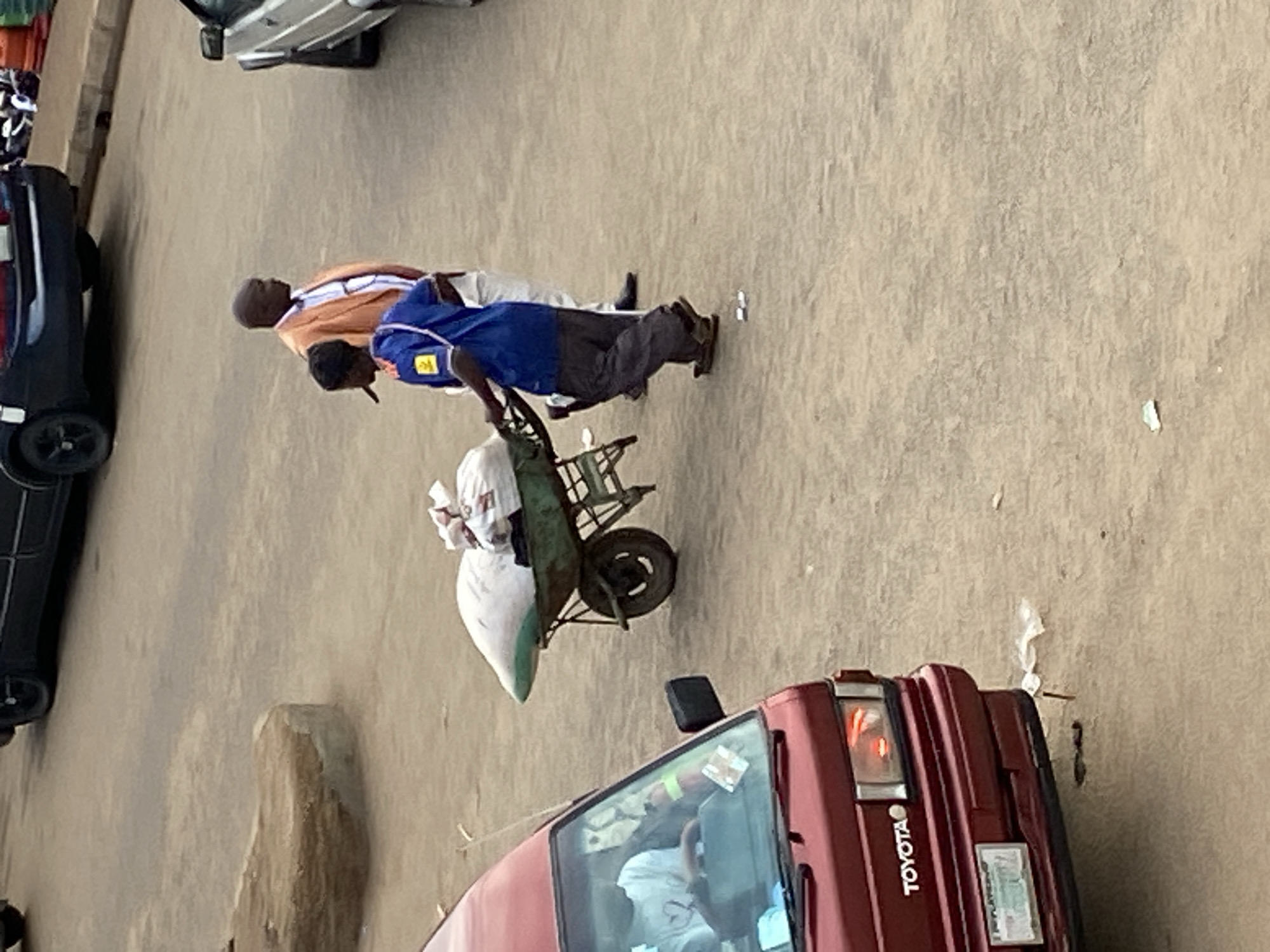 The image depicts an outdoor, dusty paved area in Bukuru, Plateau State, Nigeria, resembling a street or open market space. In the foreground-left, a maroon Toyota van is partially visible, showing its front grille with the "TOYOTA" emblem, a license plate (partially legible "PLATEAU"), and an illuminated right headlight. A person wearing a white t-shirt is seated in the driver's seat.
In the mid-ground, a person with dark skin, dressed in a blue short-sleeved shirt with yellow insignia on the left arm and shoulder, and dark trousers, is actively pushing a green metal wheelbarrow. The wheelbarrow contains two large white sacks and additional smaller items. This individual is walking towards the right side of the frame. Adjacent and slightly behind the wheelbarrow pusher, a second person with dark skin is visible, wearing an orange blazer over a white collared shirt, light-colored trousers, and dark shoes. This individual is also walking towards the right.
In the upper-left background, a dark-colored sedan is parked. In the upper-right background, the front of a silver-colored car is partially visible. A low, rounded concrete barrier or planter is situated in the mid-left background. Scattered small pieces of white paper or plastic debris are visible on the ground.