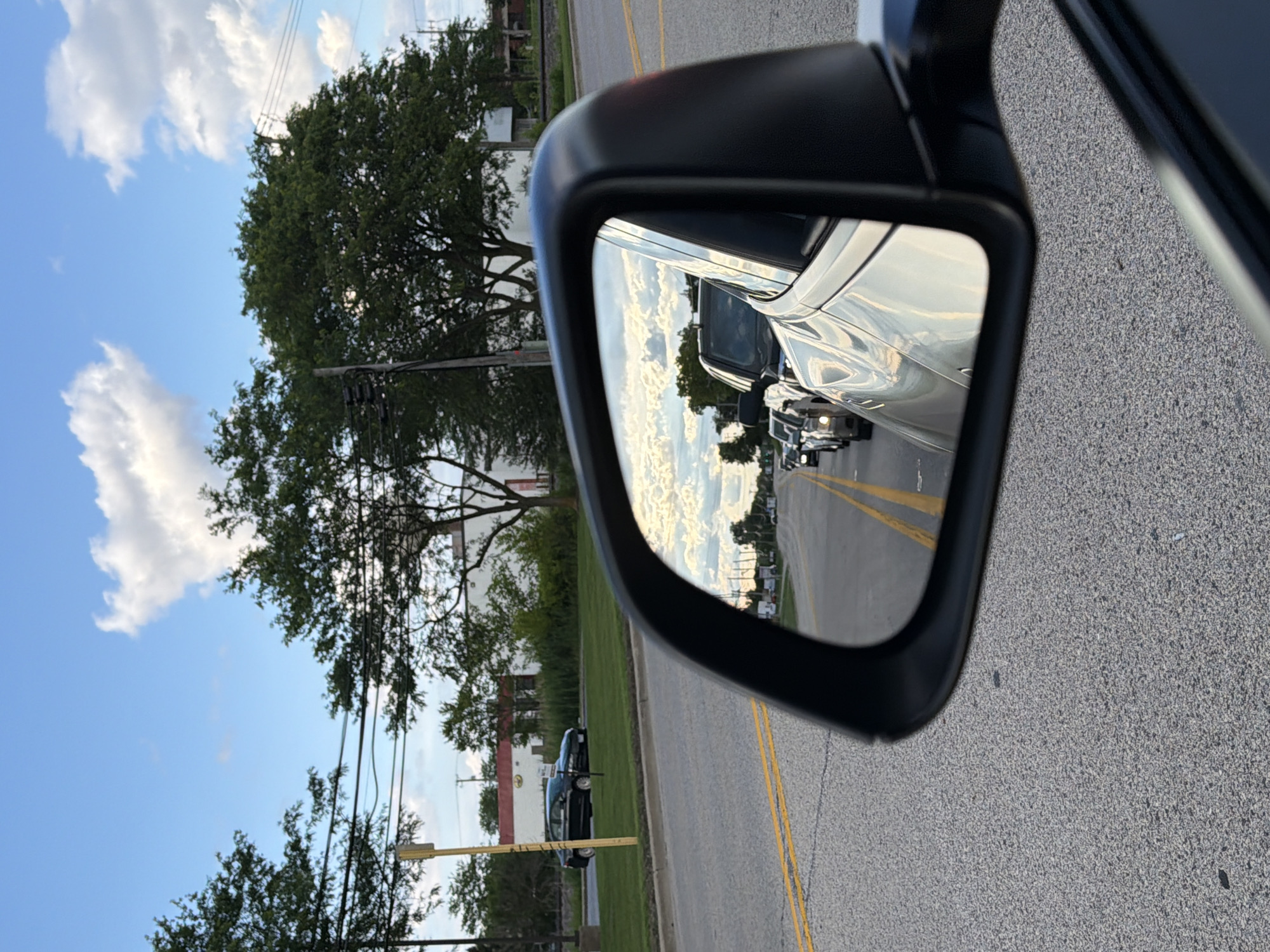The image shows a view from a car looking at the side mirror. The side mirror reflects a road with several cars in traffic, under a cloudy sky. In the foreground, the car's gray asphalt road is visible. In the background, there are trees, a building, and a street. The scene is set in Mundelein, United States of America.