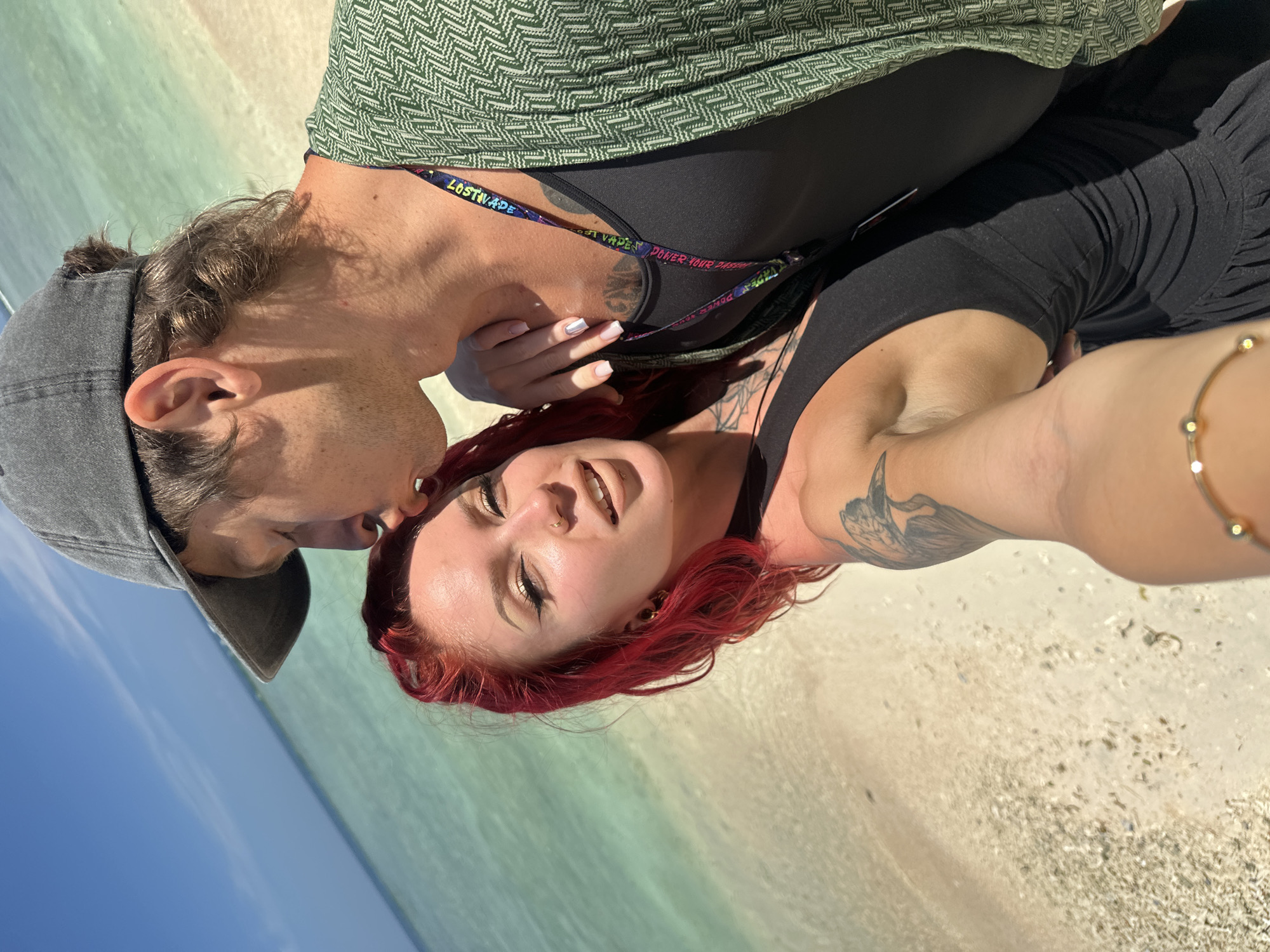The image shows two individuals in an outdoor setting, likely a beach based on the sand and water visible in the background. A person wearing a gray cap and dark clothing appears to be leaning towards another person with red hair, who is lying on the sand. The person with red hair has tattoos on their arm and chest. The background features turquoise water and a sandy beach. The lanyard has Lost Vape as one word, it says "Power Your Passion". The location is presumed to be in Unknown, Indonesia.
