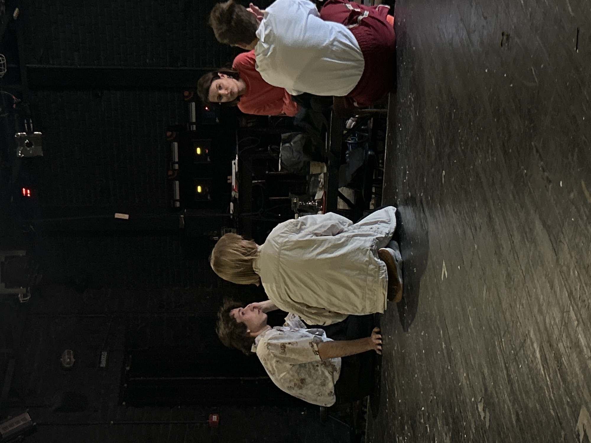 Four individuals are gathered on a dark, wooden stage. The person on the far left is a male, wearing a light-colored, patterned short-sleeved shirt and dark pants. He is sitting with his knees bent and his left hand resting on the stage floor. Next to him is a person with blonde hair, wearing a light-colored, oversized jacket and dark pants. This person is also sitting with their knees bent, facing away from the camera. To the right of them, a woman in a reddish-orange long-sleeved shirt and dark pants is seated, looking towards the person in the white shirt. The person closest to the camera on the right is wearing a white long-sleeved shirt and dark red pants with a white stripe. This person is also seated, with their back to the camera. In the background, there is a dark wall with some equipment and lighting visible. The overall scene appears to be a rehearsal or a meeting on a stage.
