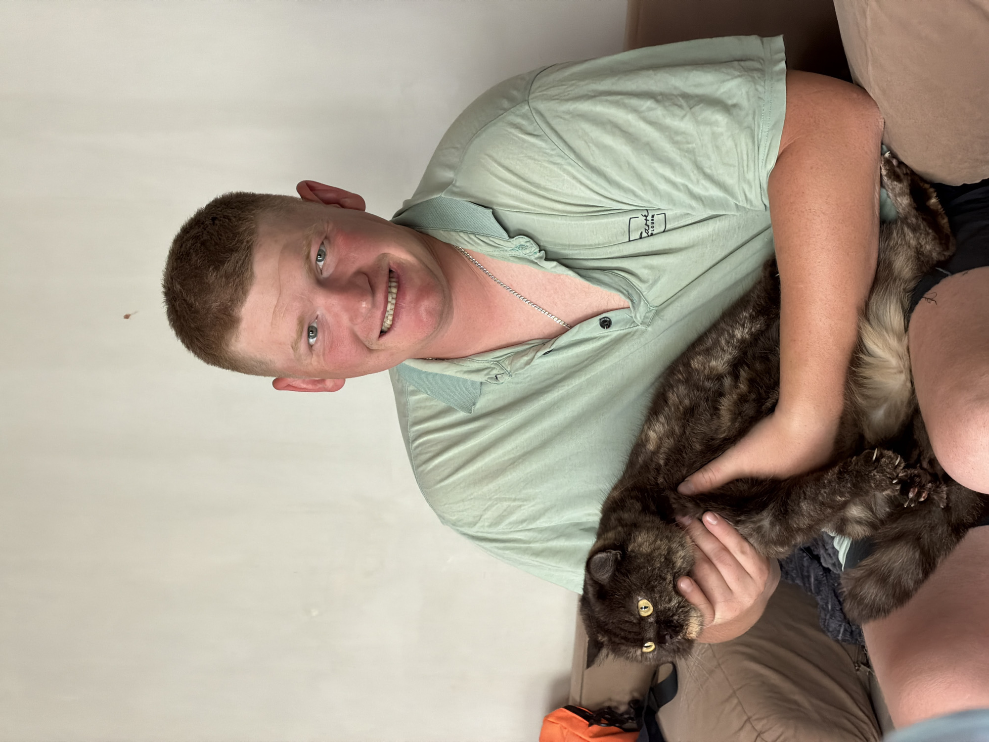 This indoor portrait, set in Chaikovsky, Russia, features a smiling man holding a cat. The image is presented as if rotated to the correct vertical orientation. The man, who appears to be young to middle-aged, has short, light brown hair and a fair complexion. He is wearing a light mint green short-sleeved polo shirt with a silver chain around his neck. His face is turned towards the camera, displaying a wide, cheerful smile that shows his teeth, and his blue-green eyes are bright.
He is gently cradling a medium-sized cat with a distinct tortoiseshell pattern, featuring a mix of dark brown, black, and reddish-tan fur. The cat has piercing yellow-gold eyes and appears calm and comfortable in the man's arms, with one of its paws showing slightly extended claws. They are seated on a light brown, upholstered couch or armchair, against a plain, light off-white wall background. A small, square logo with partially visible text, including "GLOBAL," is present on the left chest area of the man's shirt. The overall impression is one of affection and contentment between the man and his pet.